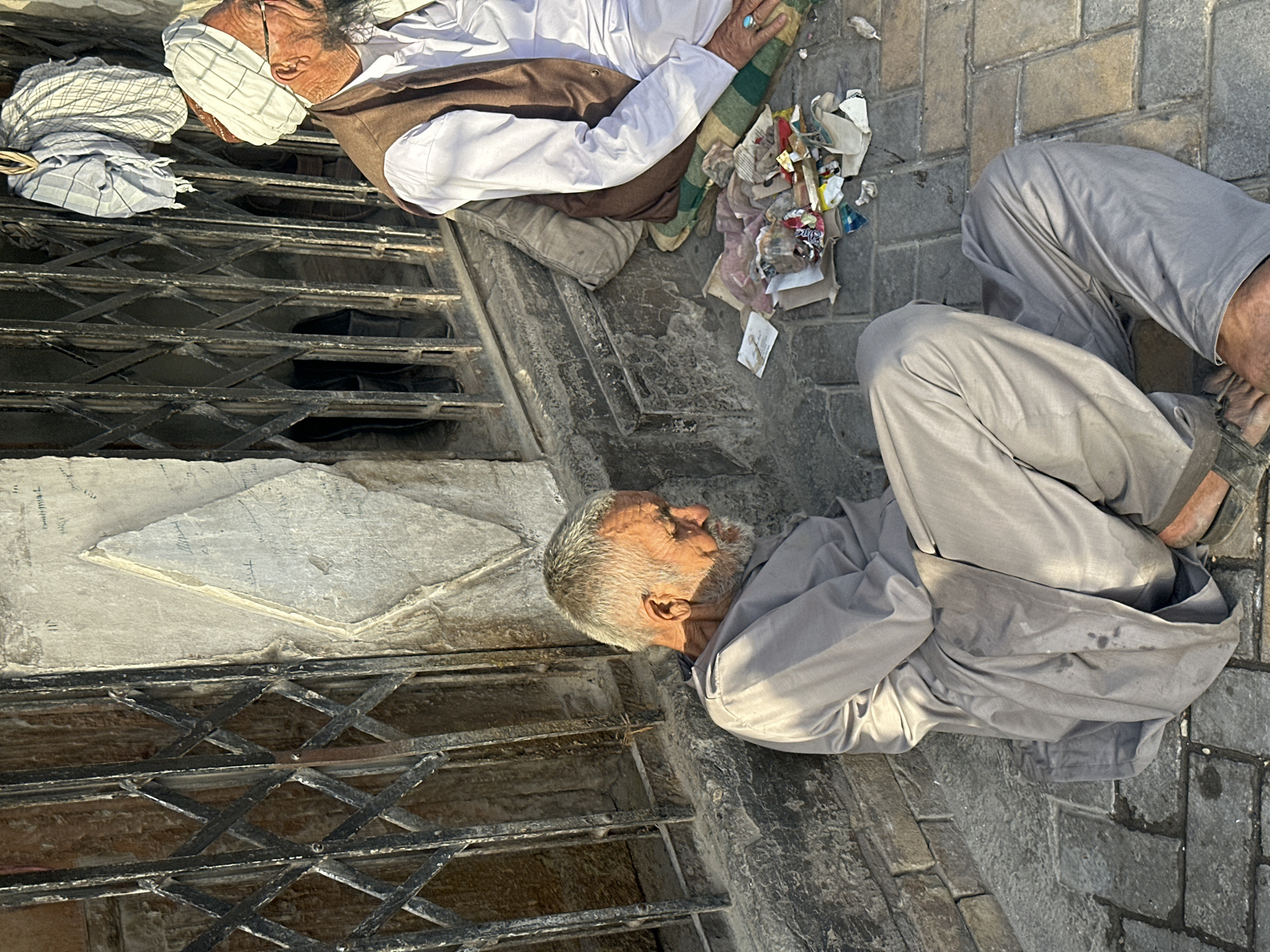 Two elderly men are depicted sitting on a paved street or alleyway in Quetta City Tehsil, Pakistan, under bright daylight conditions, evidenced by the strong shadows.
The man in the foreground, closer to the viewer, has a grey beard and is wearing light grey attire, resembling a shalwar kameez. He sits with his legs bent, facing towards the right side of the frame, appearing to be resting or waiting. His clothes show some stains or marks.
Further back and to the left, a second elderly man is seated, dressed in a white long-sleeved shirt, a brown vest, and a white and grey checkered turban covering his head. He also has a visible beard and wrinkled skin. His left hand, adorned with a ring, rests near a folded piece of greenish, striped fabric. Next to him is a small pile of miscellaneous trash, including paper scraps, plastic wrappers, and other debris, indicating a potentially neglected or public space.
Behind the men, a dark, intricate metal grate, possibly part of a storefront or entrance barrier, spans across the scene. The ground is made of grey cobblestones or pavers. To the far left, a light-colored concrete or stone structure is partially visible. No discernible text is visible in the image. The overall atmosphere suggests a moment of rest in an urban environment.