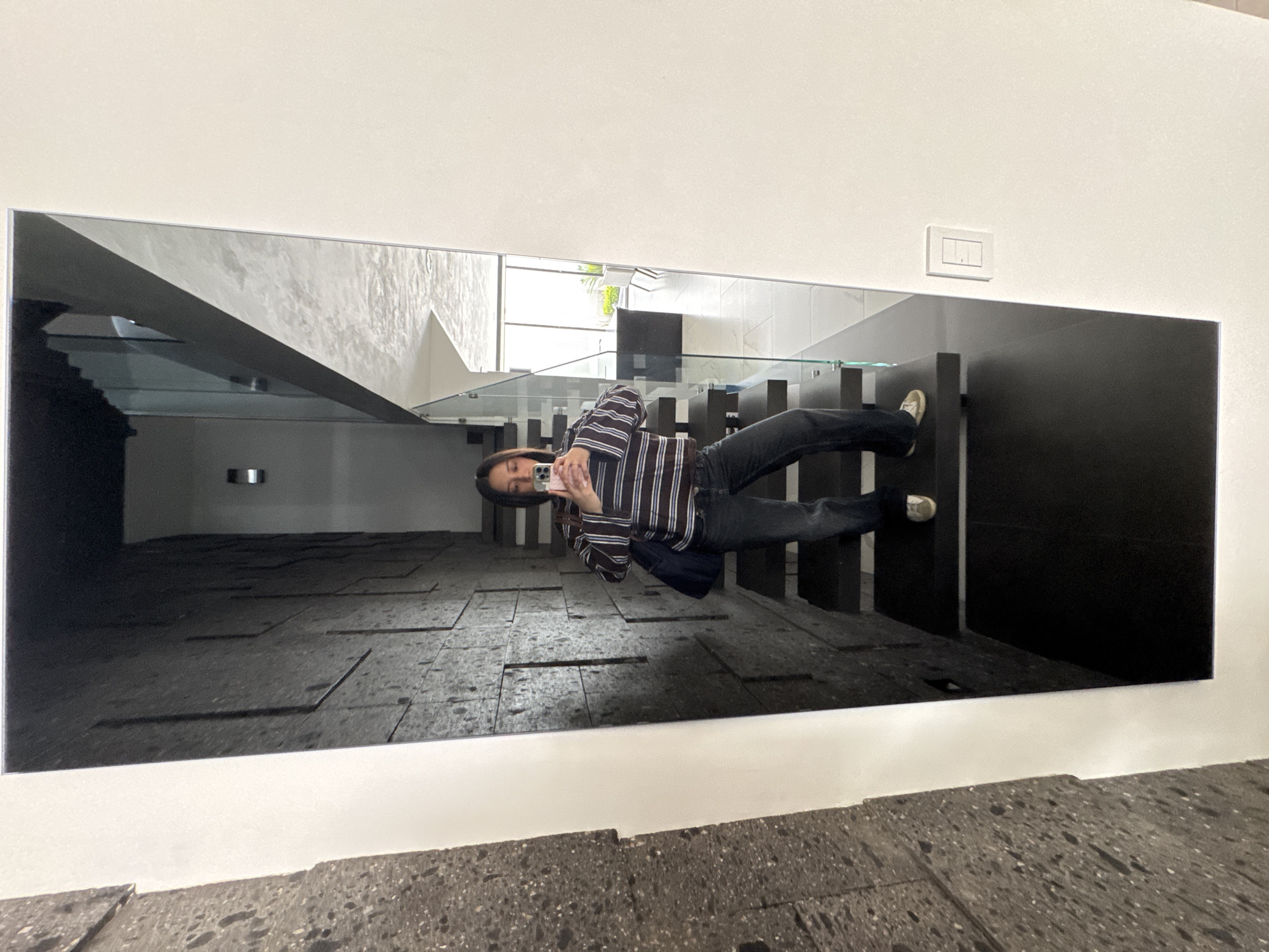 The image depicts a person taking a picture of themselves in a mirror. The person is wearing a striped shirt and dark pants. The mirror reflects an interior space with stairs, a glass railing, and a window. The location is in Puebla, Mexico. A light switch is visible on the wall above the mirror. The mirror's edge is visible, showing how it's mounted on the wall. The wall and floor beneath the mirror have a gray and white speckled pattern.