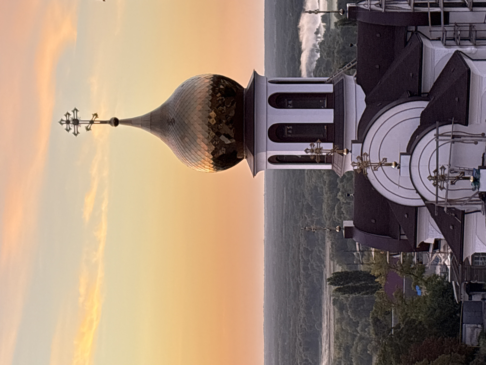 The image shows the dome and upper portion of an Eastern Orthodox church in Bobrov, Russia, during a sunset. The sky is a gradient of orange and yellow with scattered clouds. The church features a prominent golden dome, topped with a cross. Below the dome is a white tower with arched windows. The church's roof is dark brown. In the background, there is a dense forest stretching to the horizon. Patches of fog are visible in the distance, possibly over a river or low-lying land. The foreground shows more of the church's roofline, scaffolding, and some surrounding buildings, including a garage-like structure. There are no people visible in the image.