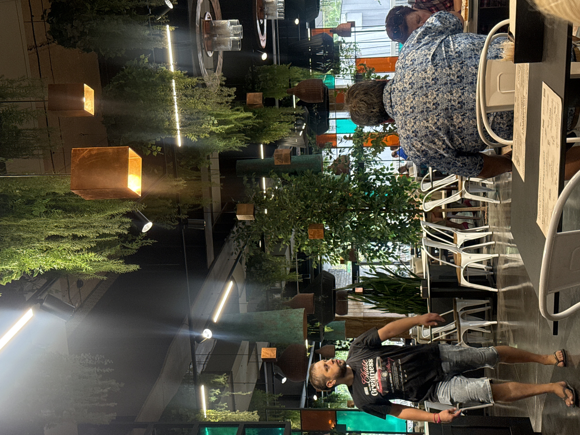 The image captures the interior of a modern restaurant with abundant hanging greenery and industrial-style lighting. In the foreground, a man with short brown hair and a beard, wearing a black t-shirt with a graphic print and light-colored denim shorts, walks towards the left of the frame. His attire suggests a casual outing. To the right, the back of another person is visible, wearing a blue and white floral patterned shirt. They are seated at a table, facing away from the camera. The environment is characterized by dark ceilings, exposed beams, and several hanging light fixtures, some of which are copper-colored and illuminate the space. Various types of chairs and tables are arranged throughout the restaurant, with some unoccupied and others occupied by patrons further in the background. The overall aesthetic is contemporary, with a blend of natural elements and urban design. The scene is well-lit, with light rays filtering through the hanging plants and from the overhead lights, creating a dynamic interplay of light and shadow. The setting appears to be a lively dining establishment.
