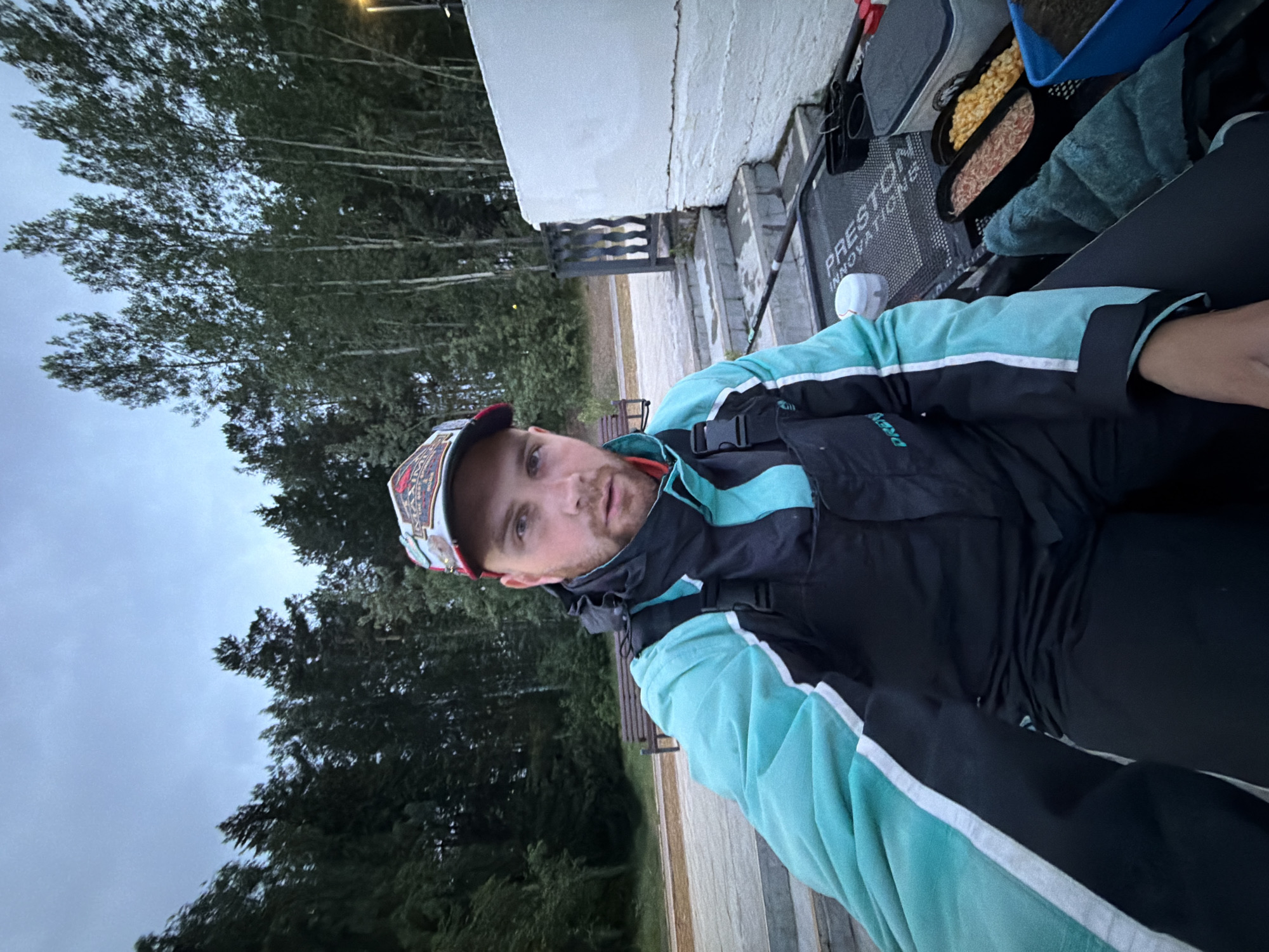 A male individual, appearing to be in his late 20s or early 30s with a light beard, is captured in a close-up, chest-up view. He is wearing a multi-colored baseball cap and a teal, black, and white jacket with "DREN" on the left sleeve, over black bib overalls. He is looking directly at the camera.
The individual is seated outdoors, positioned on what appears to be a fishing platform or seat. To his immediate right, several containers of fishing bait or food are visible on a black mesh platform, partially labeled "PRESTON INNOVATIONS." These containers include one with pink/reddish material, one with yellow corn-like kernels, and a blue container with dark brown material. A white cooler with a grey lid, a small white container, and a teal towel are also present. The handle of a fishing rod extends from the right side.
The background consists of a dense forest with tall trees, predominantly dark green, under an overcast, light grey-blue sky. To the left, a wooden platform or boardwalk is visible, leading to a wooden bench. To the right, behind the individual, there is a textured white wall constructed from blocks or stones, accompanied by steps and a dark, ornate railing. A faint light source, consistent with a streetlamp, is visible above the trees on the far right. The scene depicts an outdoor fishing setting in the city of Chaikovsky, Russia.
