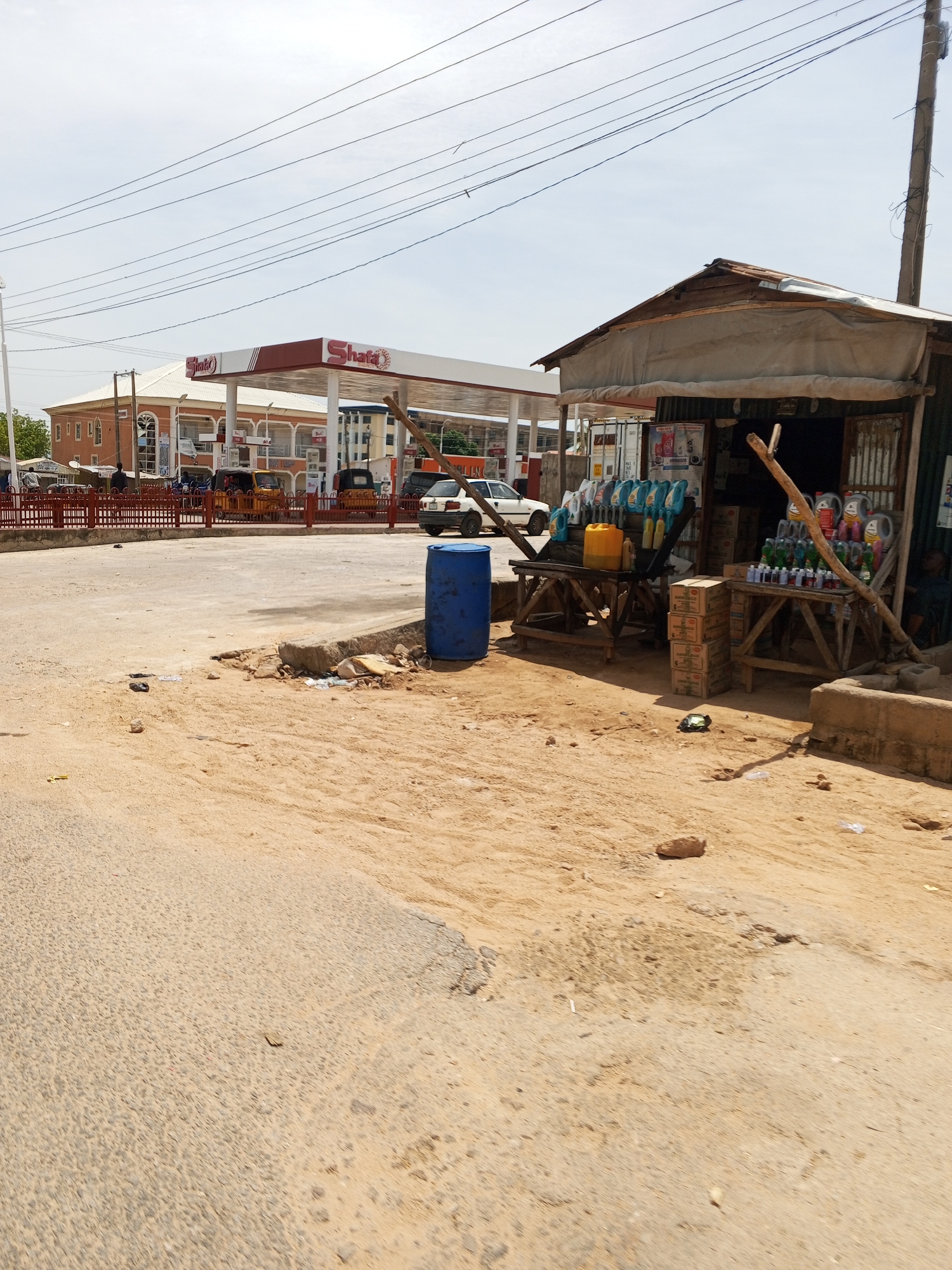 The image depicts a street scene in Jalingo, Nigeria. On the left, a Sha'a'f petrol station with its distinctive red and white canopy is visible. Adjacent to the petrol station, on the right side of the image, is a small, open-air stall constructed with corrugated metal and wood, featuring a tarpaulin roof. This stall displays various products, including containers of liquids, possibly engine oil or lubricants, arranged on wooden tables. Behind the stall, a few stacked cardboard boxes are visible. In the foreground, a sandy, unpaved area with scattered debris and rocks dominates the scene. A large blue plastic barrel is placed on the right side of the foreground. In the middle ground, a white car is parked near the petrol station, and a yellow tricycle is also present. The environment appears to be a commercial area with a mix of established businesses and informal trading setups. The sky is overcast with visible overhead power lines.