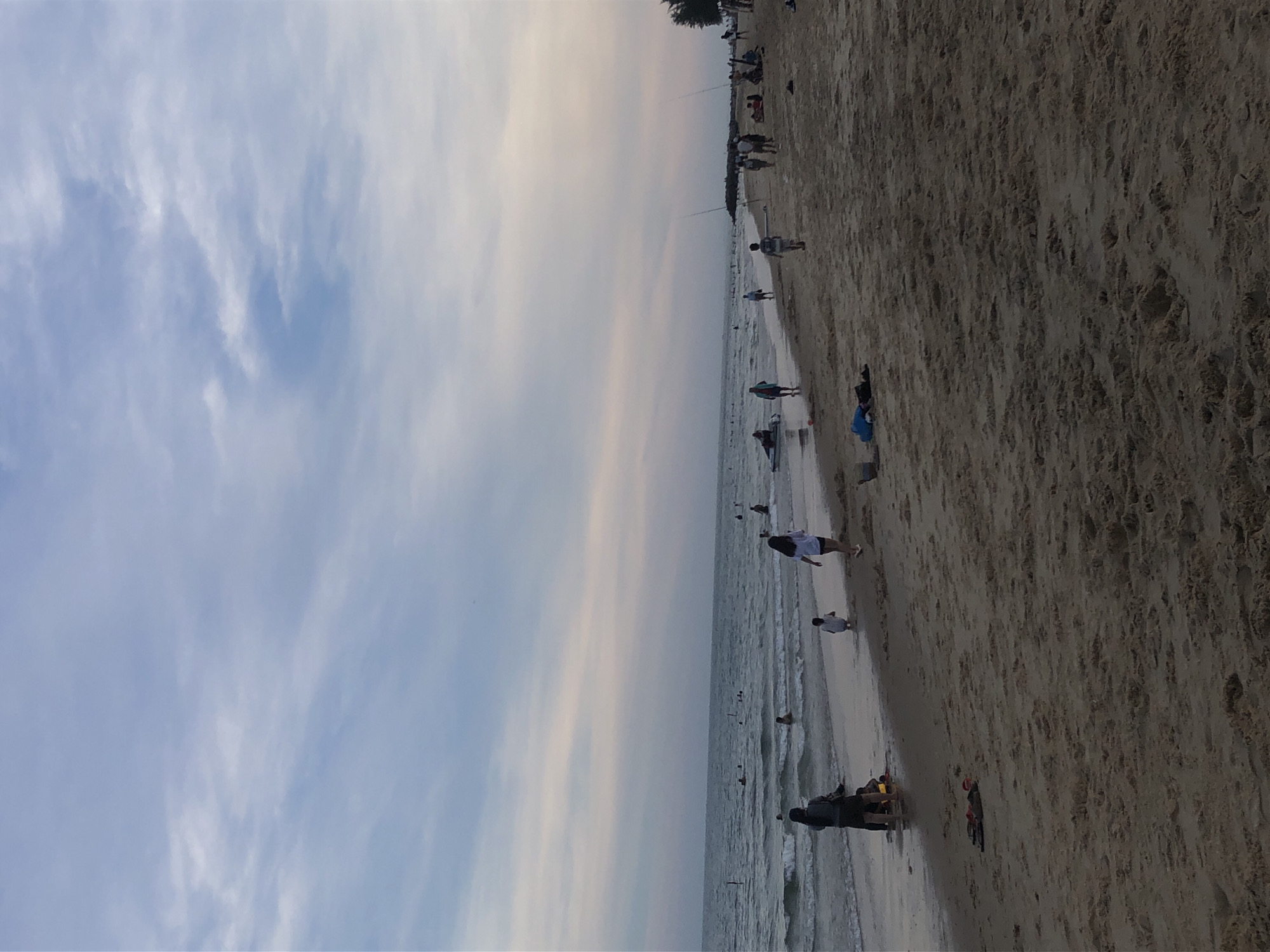 The image depicts a beach scene in Mũi Né, Vietnam, likely during the late afternoon or early evening given the soft light and pastel colors in the sky. The sky is a blend of light blue and pale pink, with scattered clouds. The ocean is calm with gentle waves rolling onto the shore.
Several people are visible on the beach and in the water. On the shore in the foreground, a woman in a dark top and shorts is walking towards the right, and a child in a white outfit is crouching near her. Further down the beach, a group of people are gathered, some sitting and others standing. In the water, multiple individuals are swimming or wading, and a person is on a small, dark-colored watercraft.
The sand on the beach is a light brown color and appears somewhat textured, with some footprints and debris scattered across it. Along the waterline, small waves are breaking. In the background, a line of tall, thin poles, possibly fishing rods or markers, are visible stretching along the shore. The overall atmosphere is peaceful and relaxed, indicative of a typical beach day.