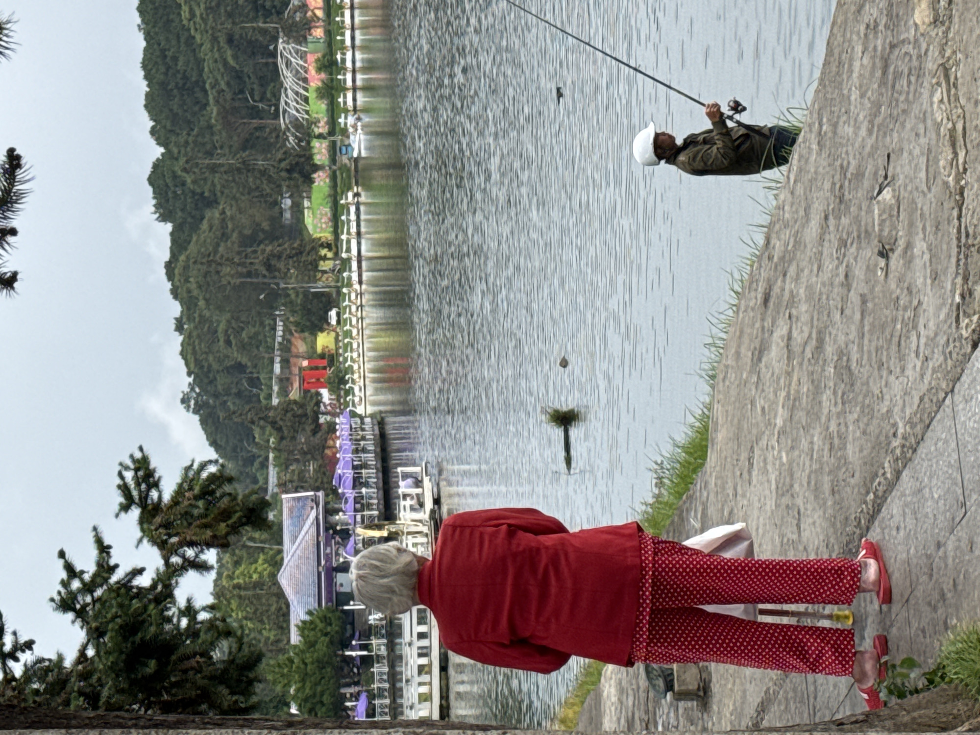 The image depicts Xuan Huong Lake in Da Lat, Vietnam, on an overcast day. In the foreground, an elderly woman with grey hair, facing away from the camera, stands on a paved walkway. She is wearing a red blazer and red polka dot pants, with red slippers on her feet. She holds a white bag in her left hand. To the right, a person wearing a white helmet, a dark jacket, and dark pants is fishing in the lake with a rod. The lake is calm with ripples on its surface. In the background, across the lake, a row of white swan paddle boats are visible near a waterfront promenade lined with purple umbrellas and various buildings, some with red accents. Dense green trees and vegetation cover the hillsides beyond the lake.