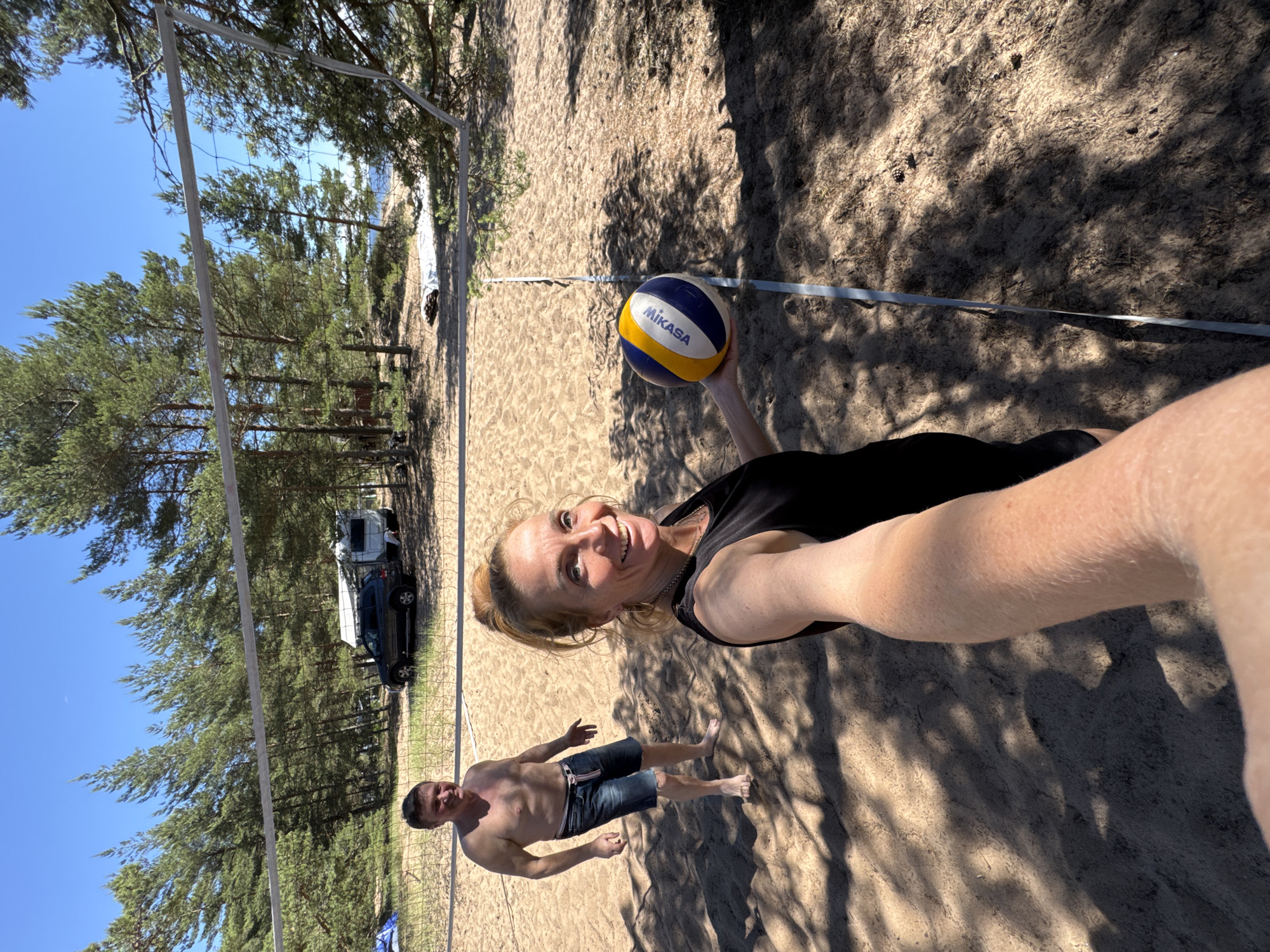 A middle-aged Caucasian woman with blonde hair pulled back in a bun, wearing a black sleeveless top, is holding a volleyball and taking a selfie on a sandy beach. She is smiling and looking at the camera. Behind her, a shirtless middle-aged Caucasian man with short dark hair is standing in a volleyball court. He is wearing denim shorts and is looking towards the camera with his arms slightly out. A volleyball net is strung across the court between them. In the background, there are pine trees, a car, and a small caravan. The setting appears to be a beach or a campsite. The image appears to have been taken on a sunny day. There is no specific indication that this location is in Zelenaya Roshcha, Russia, or any other specific city.