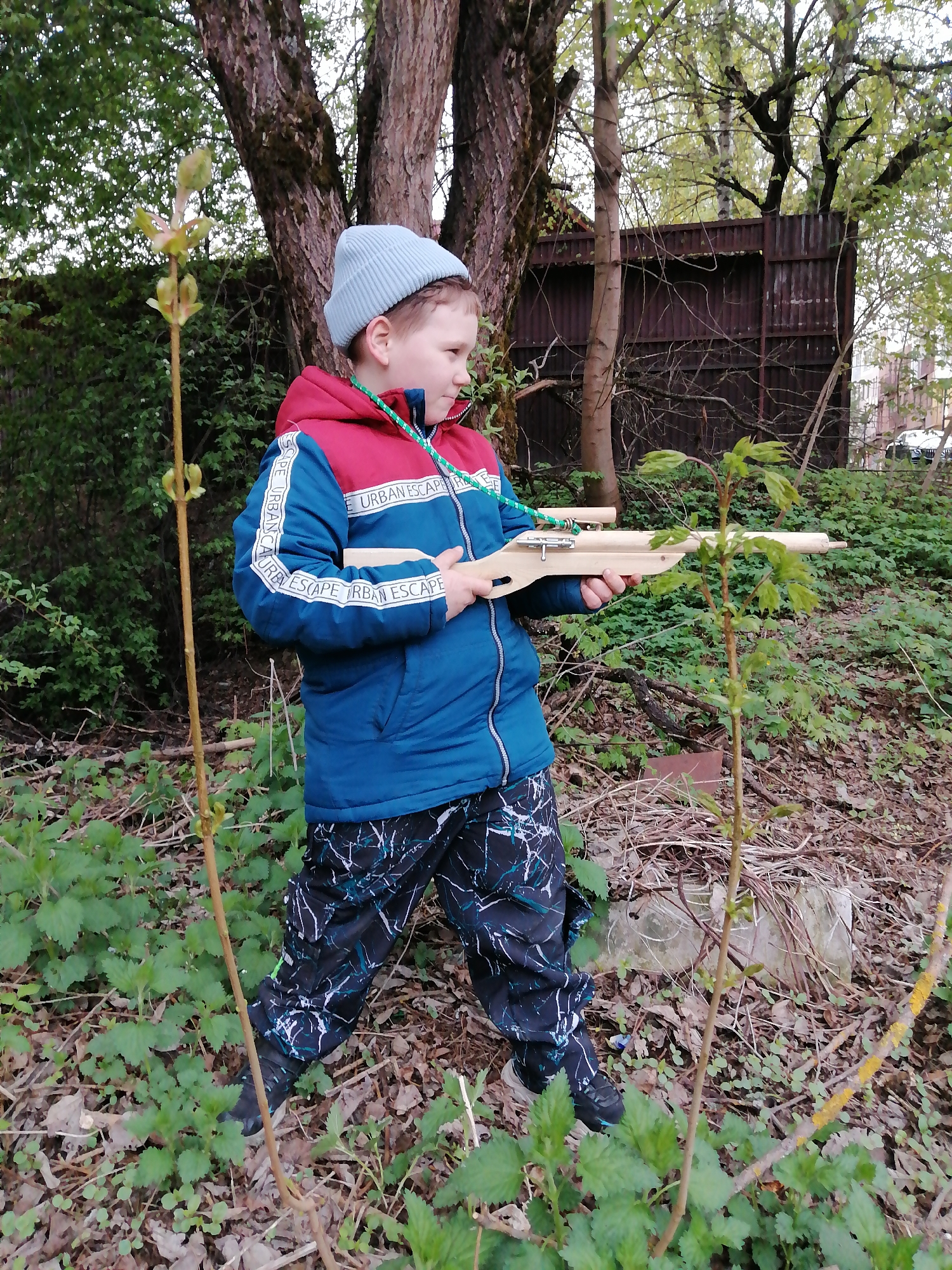 A young boy stands in a wooded area with dense ground cover, holding a wooden toy crossbow. He wears a blue beanie, a blue and red jacket with "URBAN ESCAPE" printed on the sleeves, and black patterned pants. The backdrop features trees, bushes, and a dark wooden fence in the distance. The location is the city of Unknown, Unknown.