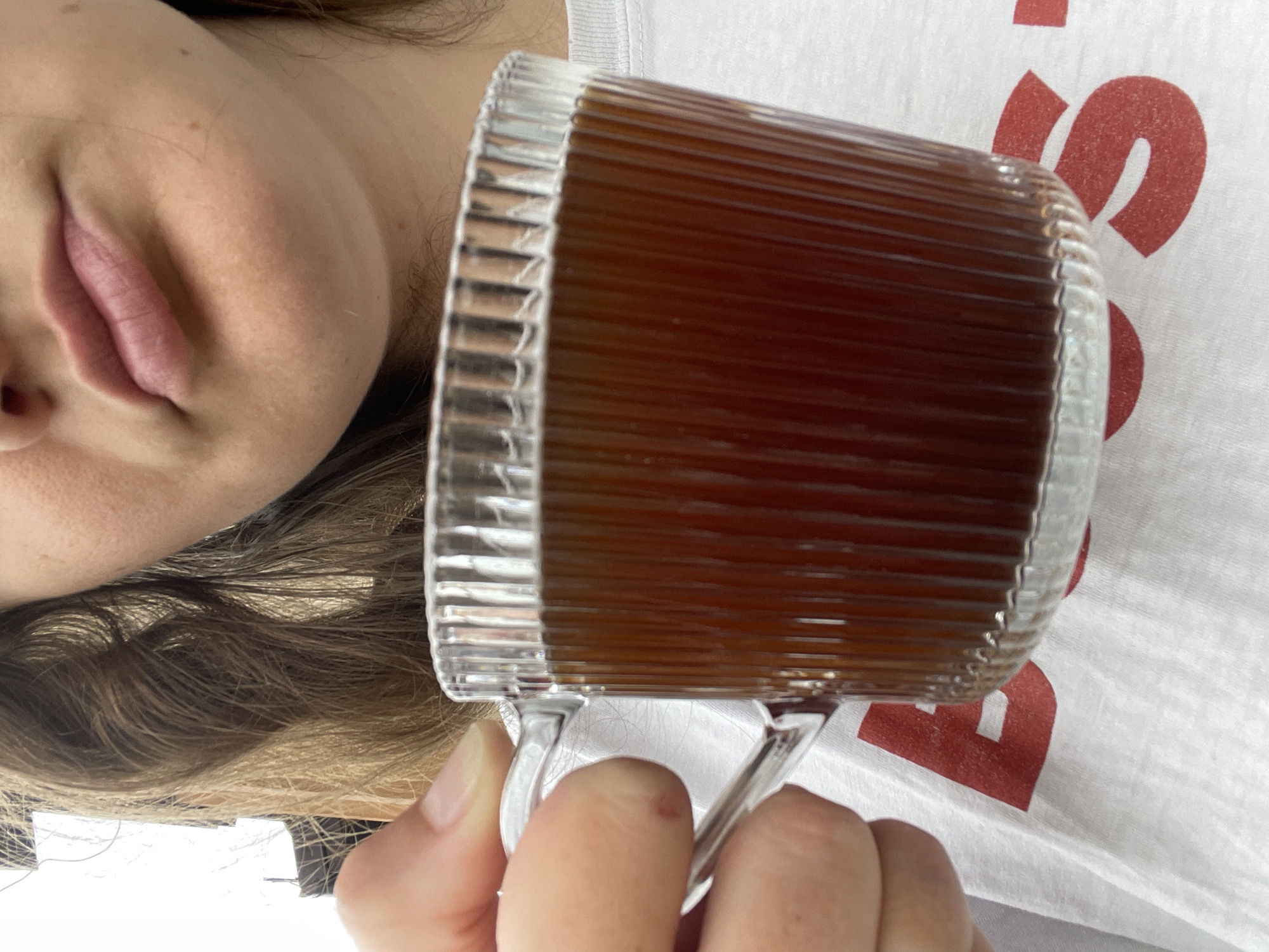 A close-up, medium shot captures a person holding a clear, fluted glass mug filled with dark liquid, presumably coffee, against a blurred background. The mug has a ribbed texture and a clear glass handle. The person's fingers, with light-colored nail polish, are visible gripping the handle, and a small reddish mark is present on the thumb. The person's mouth, with slightly parted lips and a light sheen, is visible in the upper portion of the frame, suggesting they are about to take a sip or have just taken one. They are wearing a white t-shirt with a large red letter "B" printed on it. The background is out of focus, with hints of brown hair and bright white light, suggesting an indoor setting with diffused lighting. The overall scene is casual and intimate, focusing on the act of enjoying a beverage.