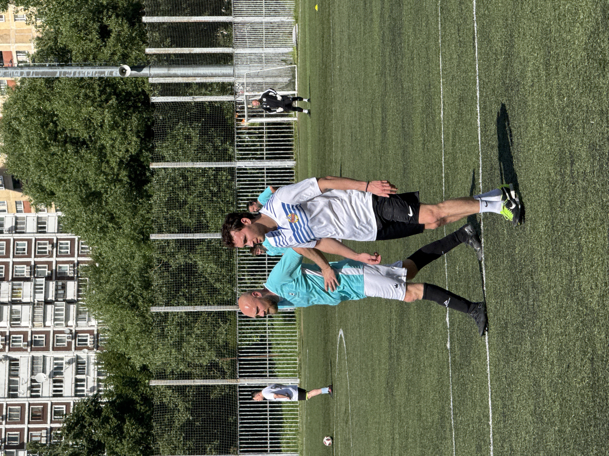 A group of men are on a green turf soccer field on a sunny day. In the foreground, a man in a white and blue striped soccer jersey and black shorts is walking. To his left, a bald man in a turquoise soccer jersey and white shorts is walking. In the background, another man in a white soccer jersey and black shorts is standing near a soccer ball. Further back, a man in a black and white goalkeeper uniform stands in front of a soccer goal. Behind the field, there is a chain-link fence and lush green trees. In the far background, a tall, multi-story apartment building with numerous windows is visible.