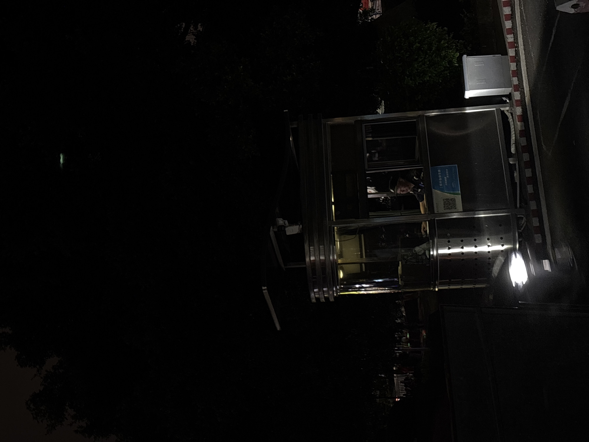The image captures a nighttime outdoor scene in Nan'an District, China, centered around a brightly lit, metallic booth.
**Subjects:** Inside the booth, a person is visible, their face partially illuminated. They appear to be wearing a dark uniform, possibly a cap, and are positioned as if looking outwards or working within the booth.
**Objects:** The main object is a modern, rectangular booth constructed with metallic panels and large glass windows. On its roof, a silver-colored, vented rectangular unit, likely an air conditioning component or electrical housing, is situated. To the right of the booth, a bright, modern light fixture provides significant illumination to the immediate area. Adjacent to the booth, a curb with distinctive red and white painted stripes demarcates what appears to be the edge of a dark asphalt road or pathway. Dark foliage from trees or bushes can be seen in the background and on the left side of the frame, contrasting with the artificial lighting.
**Scene and Setting:** The setting is clearly nighttime, evidenced by the darkness and the reliance on artificial lights. The booth, positioned by what seems to be a roadside, suggests a potential guard post, security checkpoint, or a small service kiosk. The overall environment is calm, illuminated only by the booth's internal lights and the external lamp.
**Activity or event taking place:** The primary activity depicted is a person on duty inside the booth, likely performing surveillance, administrative tasks, or providing service. The scene is static, showing a moment of routine rather than a specific event.
**Notable details:** The dominant feature is the artificial lighting cutting through the darkness, highlighting the booth and its immediate surroundings. The red and white striped curb is a common road marking.
**Visible text:** Inside the booth, on a blue sign, white Chinese characters are visible. The main text appears to read "倡导健康生活 | 共建美好家园" (Advocate for healthy living | Build a beautiful home together), accompanied by smaller text and a prominent QR code below.