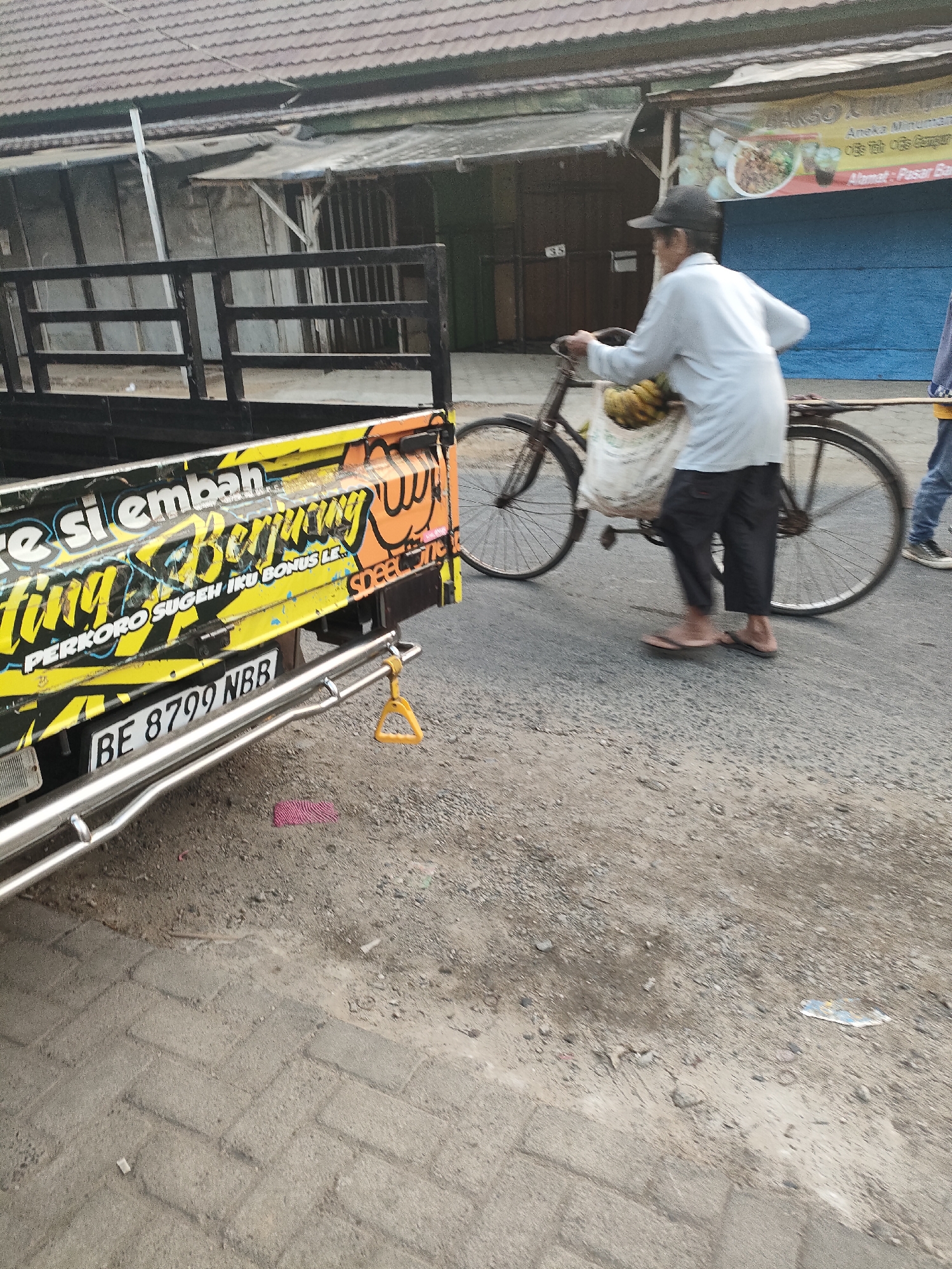 The image shows a man on a bicycle on a road in Desa Banyumas, Indonesia. The man is wearing a white shirt and black pants, and he is carrying a bag of bananas on the front of his bicycle. The bicycle has a rack on the back, and there is a reflection of the man and the bicycle in the rearview mirror of a parked truck. The truck is yellow and has a license plate that reads "BE 8799 NBB". The truck also has some graffiti on the side that says "Si Jembah" and "Beruang". The background of the image shows a building with a tiled roof. The road is made of asphalt, and there are some pebbles and debris on the side of the road. The sky is overcast.