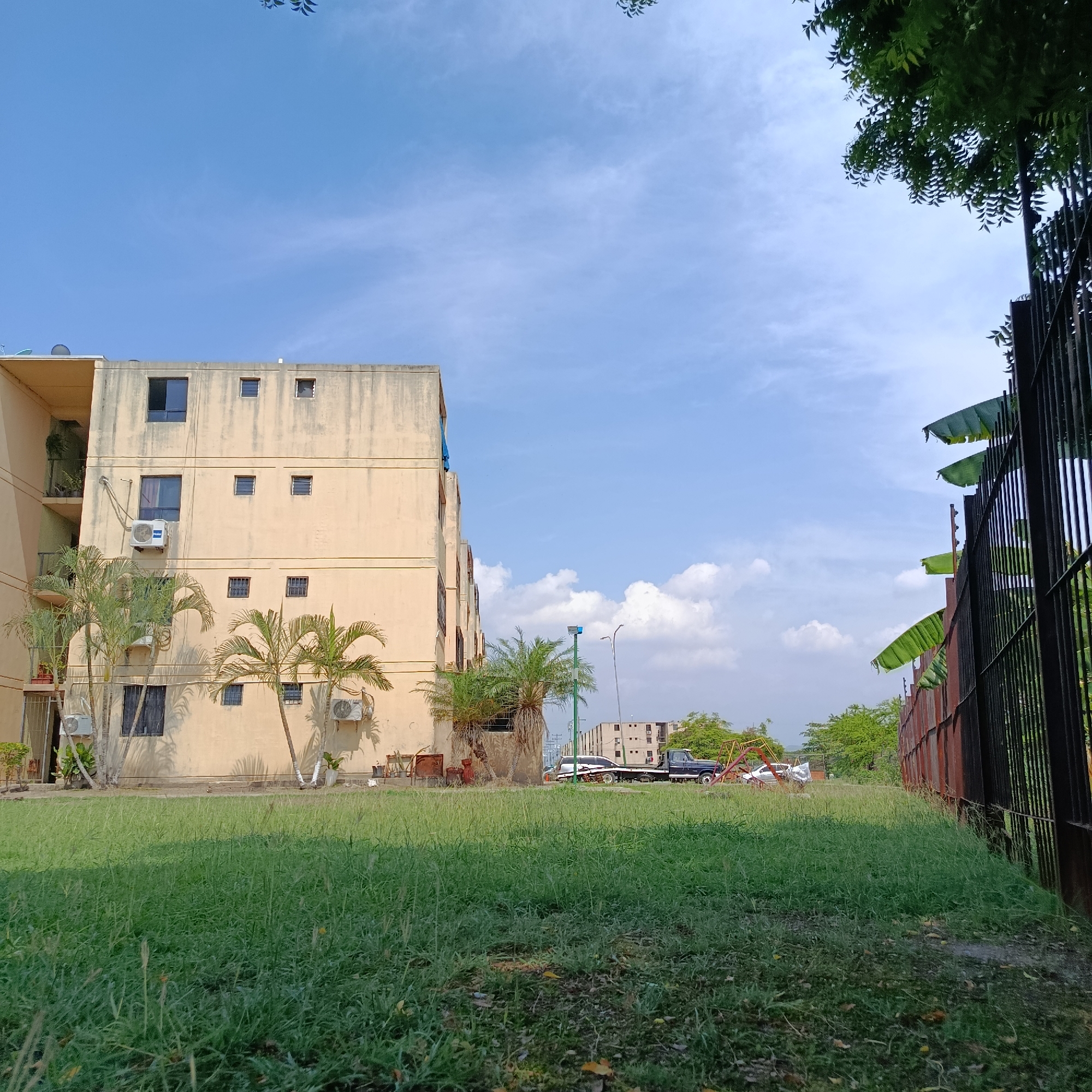 The image depicts a residential area in Los Guayos, Venezuela. A light yellow, multi-story building with multiple air conditioning units attached to its facade stands to the left of the frame. Palm trees are planted around the building's base. In front of the building is a grassy lawn. Beyond the lawn, a flatbed truck is parked, alongside what appears to be abandoned playground equipment. A tall, black metal fence runs along the right side of the frame, with banana tree leaves extending from behind it. The sky is blue with scattered white clouds.