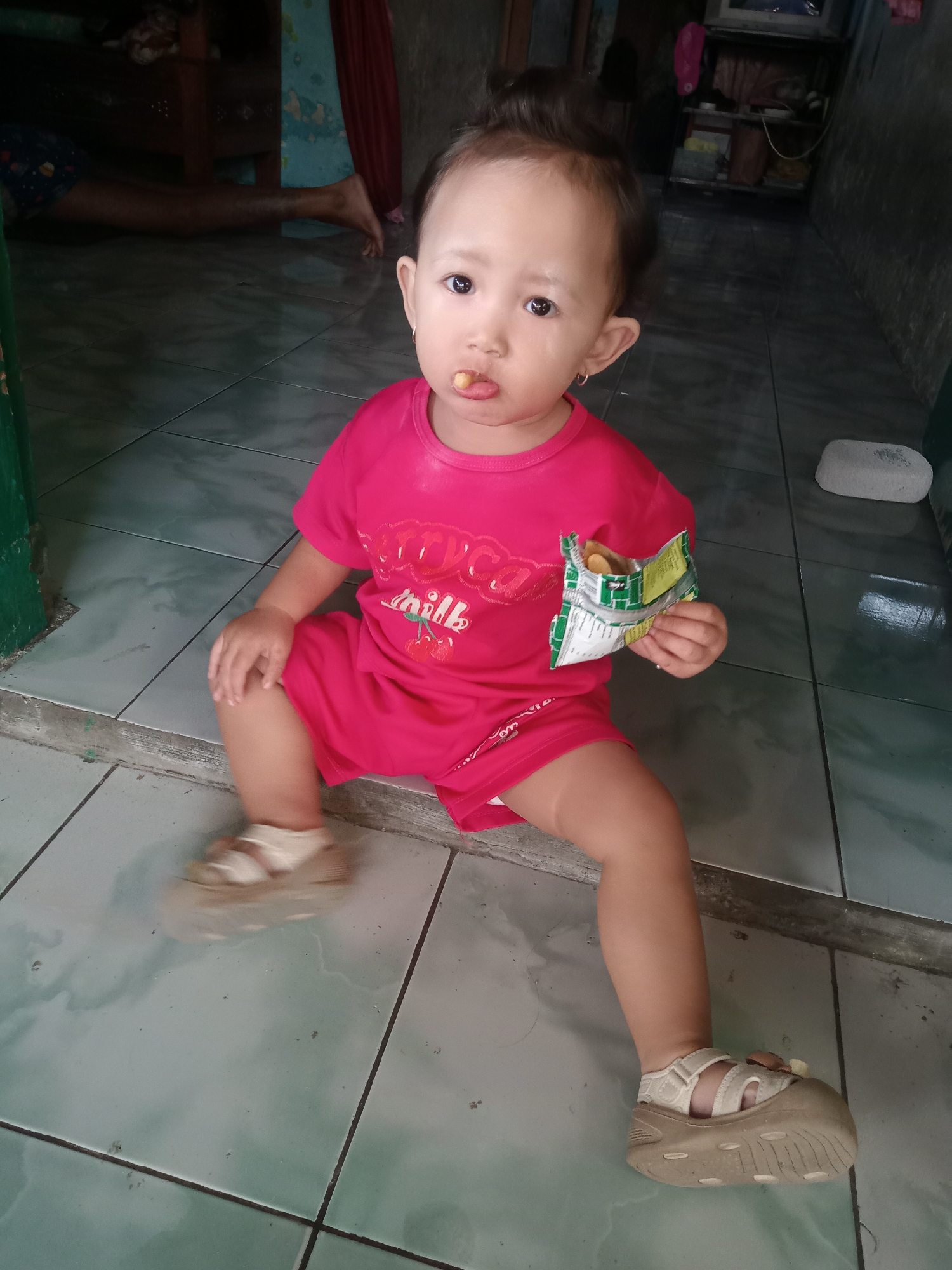 A toddler is sitting on a tiled floor, wearing a red shirt and shorts, and beige sandals. They have a snack in their mouth and hold a partially opened packet of chips in their right hand. In the background, there are hints of a living area with furniture and a person's legs visible. The scene appears to be in a home setting, consistent with the city of Bungur, Indonesia. The floor has large square tiles.
