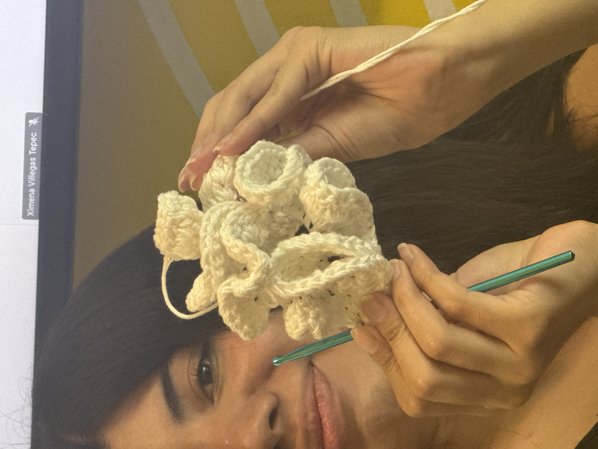 A close-up, rotated portrait image captures a woman engrossed in crocheting indoors. The image is oriented sideways, with the woman's head on the left and her body extending to the right. Her hands are central, skillfully manipulating a cream or off-white crocheted piece and a teal-colored crochet hook. The unfinished crochet work has a distinctive ruffled, organic texture, resembling coral or a cloud formation, with a strand of yarn extending from it.
The woman has dark hair and her face is partially visible; her right eye, part of her nose, and a slight smile on her lips suggest focus and contentment. Her hands are fair-skinned, with neatly manicured nails.
The setting appears to be a casual indoor environment. To the left, a portion of a dark screen or monitor is visible, featuring a vertical label or tab. In the upper right quadrant of the background, a pattern of alternating light yellow and white stripes can be seen, possibly from a wall, curtain, or blanket. The rest of the background is a darker, blurry surface, likely clothing or furniture.
The visible text on the screen label reads "Ximena Villegas Tepec," accompanied by a small icon, suggesting a user profile, document, or video title. Given the provided context, this activity likely takes place in Mexico City, Mexico, possibly in a home setting. The time of day appears to be during daylight hours or under bright artificial light.