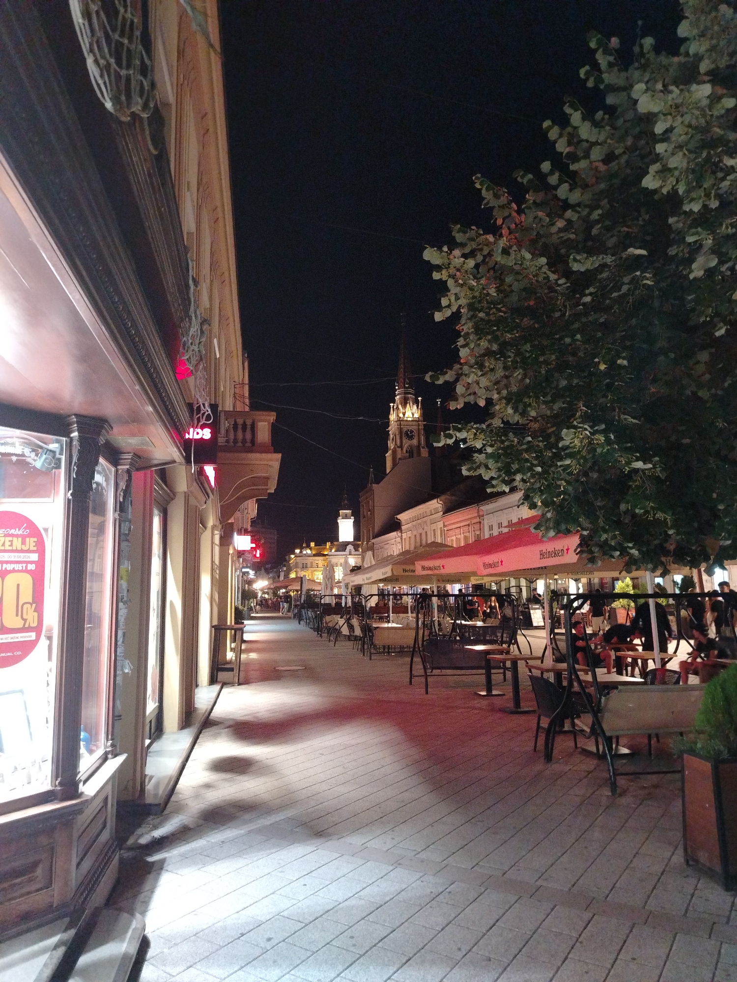 The image depicts a night scene on a pedestrian street in Novi Sad, Serbia, likely Zmaj Jovina street, known for its shops and cafes. On the left, a shop window displays a large sign with red text and a graphic of a percentage discount. The building facade above the shop is ornate, with wooden trim and decorative elements. Red neon lights and hanging string lights illuminate the street.
The street is paved with light-colored cobblestones. Along the right side of the street, several outdoor seating areas for cafes are visible, featuring tables, chairs, and red awnings bearing the "Heineken" logo. Some of these areas are empty, while others have patrons seated. A large tree with green foliage stands prominently on the right side of the image, its branches extending over the street.
In the background, the architecture of buildings lining the street is visible, with some featuring balconies and decorative facades. Further down the street, a church tower with a lit clock face rises against the dark night sky, likely the Roman Catholic Cathedral of the Name of Mary. Several people are walking along the street and seated at the cafes. The overall atmosphere is that of a lively, illuminated urban evening.