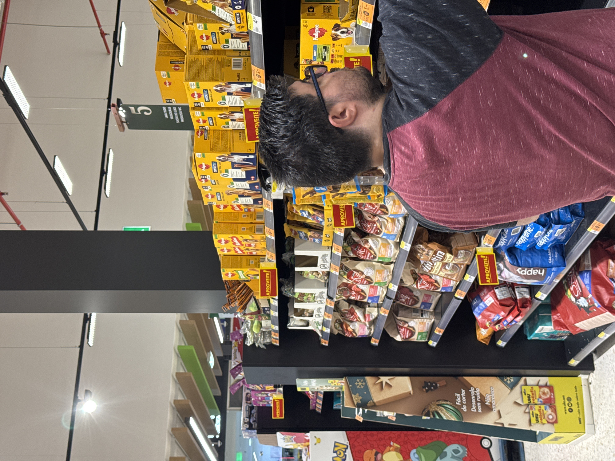 The image shows a man in a maroon and black raglan shirt browsing the pet food aisle of a supermarket. He is wearing glasses and has short, dark hair. The shelves are stocked with various brands and sizes of dog food bags and boxes, some with prominent "Aproveite!" (Take advantage!) signs. A large black pillar divides the aisle. The supermarket lighting is bright, with visible fluorescent fixtures and some natural light entering from unseen sources. In the background, there are more shelves with products, including a display with Pokémon-themed packaging on the left. The overall environment suggests a commercial retail setting, likely a large supermarket chain. The specific location of Castelo Branco, Portugal, is not visually discernible from the image content itself; it is provided as context outside the visual information.