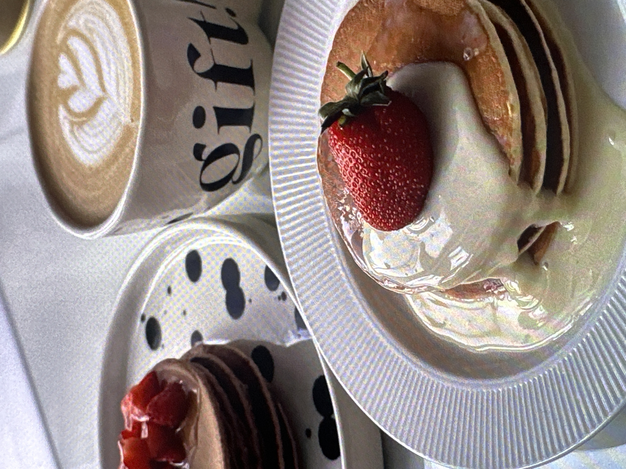 The image depicts a breakfast scene indoors. A white ceramic mug with a cappuccino, featuring latte art, sits next to a plate of pancakes topped with a strawberry and cream. A second plate with chocolate pancakes and strawberries is also visible. The plates appear to be textured, and the mugs are decorated with the word "gift!" in black lettering and a pattern of black spots. The scene is likely set in a cafe or restaurant. There is no evidence to confirm the location in Yerevan, Armenia. The image contains no visible people or interactions.