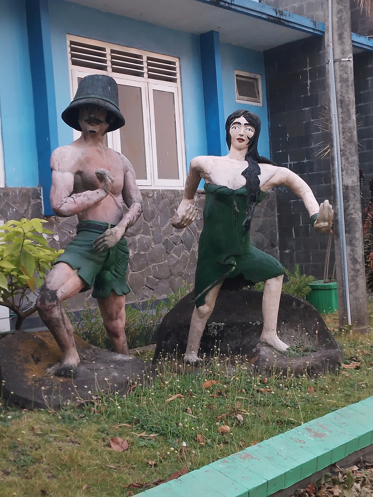 This image depicts two life-sized, weathered statues in an outdoor setting, likely in the city of Patianrowo, Indonesia. The statue on the left is a male figure wearing a wide-brimmed hat and green shorts. He has a muscular physique and is posed with one arm bent and his fist clenched. The statue on the right is a female figure wearing a green, draped garment. She has long black hair and is posed with her arms extended, one hand held up with fingers spread. Both statues appear to be made of a stone-like material and are positioned on rocks in a grassy area. Behind the statues, there is a building with blue and grey walls, featuring windows and a stone facade. A green planter box and some vegetation are visible in the background. The ground is covered in grass, scattered leaves, and small white flowers. A bright green border runs along the bottom edge of the image. The overall scene suggests an outdoor decorative element or memorial.