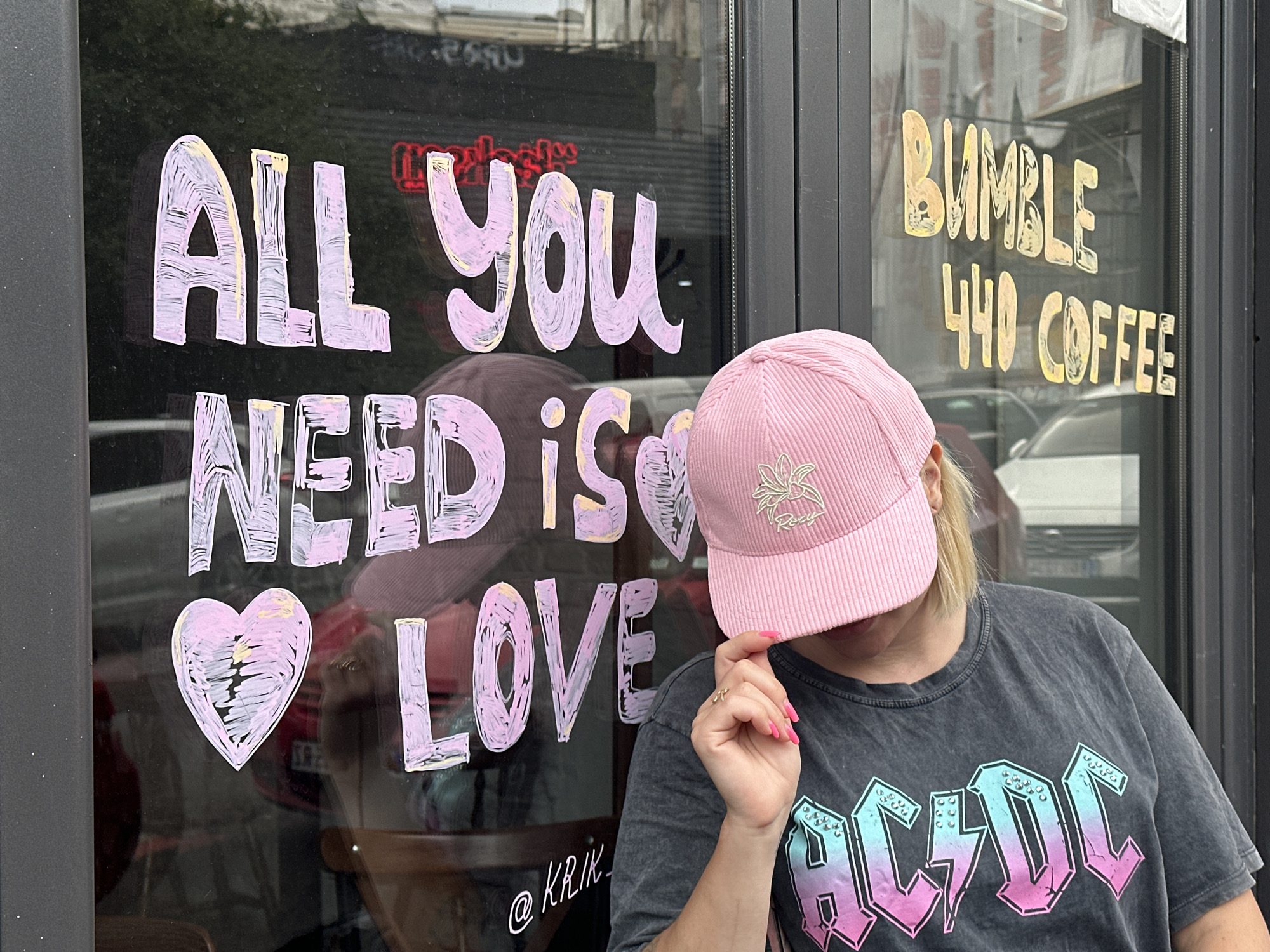 A blonde individual wearing a pink corduroy baseball cap and a dark grey AC/DC t-shirt is positioned in front of a shop window in Belgrade, Serbia. The window displays chalk art with the text "ALL YOU NEED IS LOVE" in pastel colors, including pink and yellow. To the right, the shop's name, "BUMBLE 440 COFFEE," is also written in chalk in yellow and white. The individual is partially obscuring the text, holding the brim of their cap with their right hand, which has bright pink fingernails and a delicate ring. A heart symbol is drawn on the window to the left of the word "LOVE." The reflection in the window shows a street scene with cars and buildings. The overall atmosphere is casual and friendly.