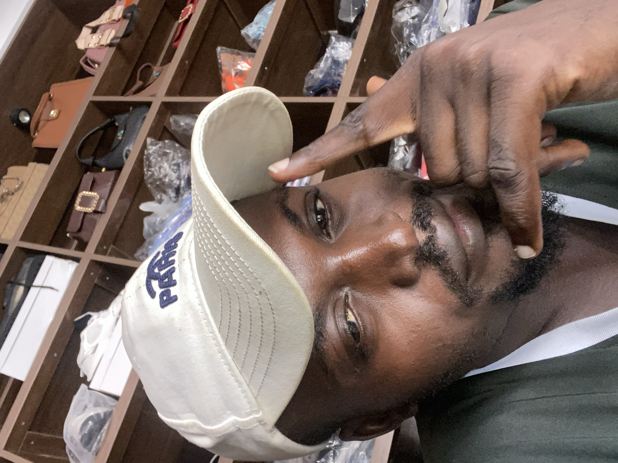 An overhead, close-up, selfie-style shot shows a Black man wearing a white baseball cap and a dark green shirt. He is looking at the camera, and his right hand is raised to his face, with his index finger touching his forehead. The background is a wooden shelving unit filled with various items, including handbags, shoes, and other packaged goods. The man has a goatee and a mustache. The image appears to have been taken indoors, possibly in a retail environment.