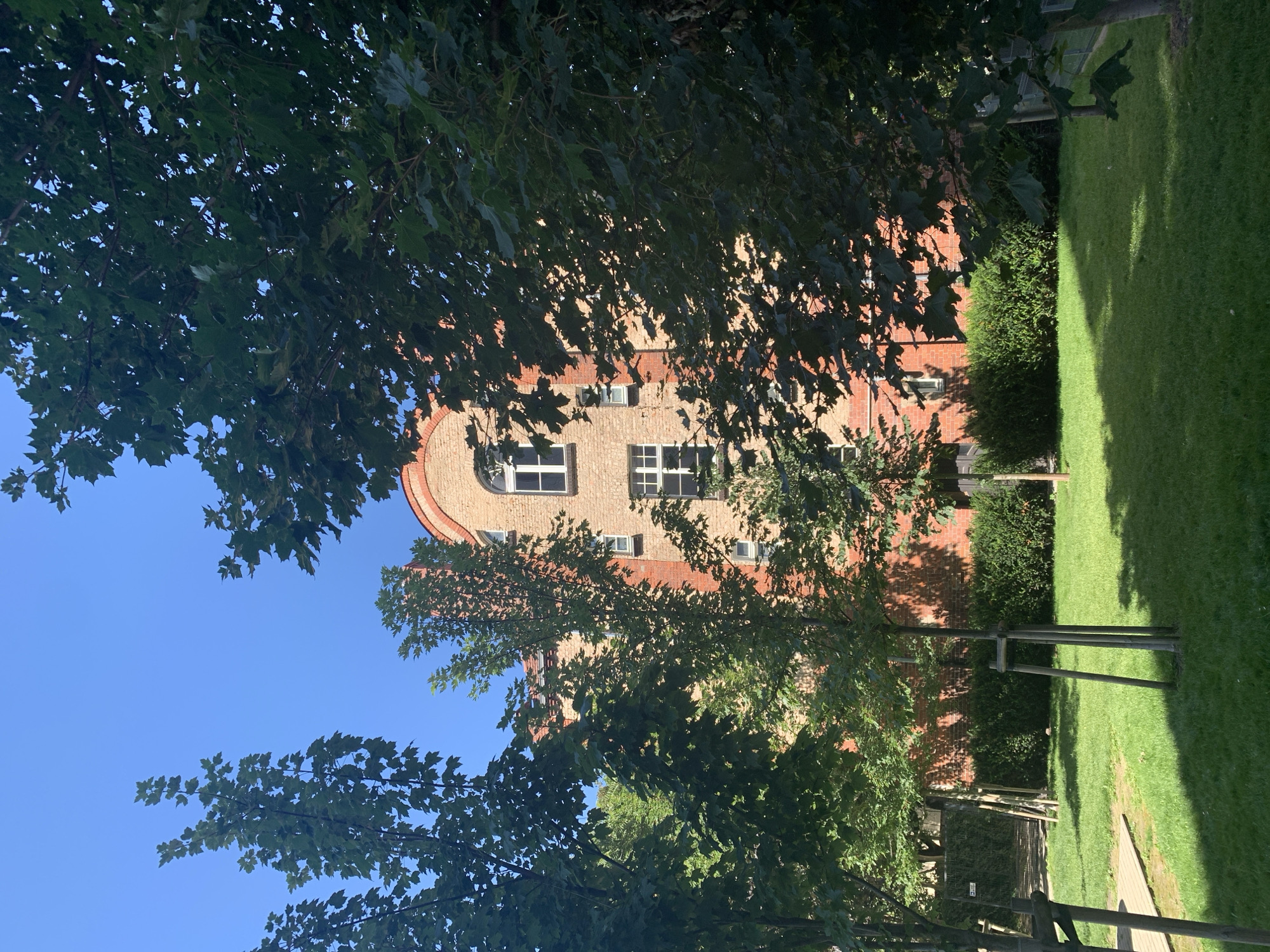 The image depicts a brick building in Poznan, Poland, partially obscured by lush green trees. The building features arched windows with white frames and is constructed from light orange bricks, with some reddish-brown brick accents around the windows and at the top corners. A curved red brick section crowns the building on the right side. In front of the building, a well-maintained lawn is visible, dotted with young trees staked with wooden poles. The sky is a clear, bright blue. The overall scene suggests a peaceful outdoor setting in a city.