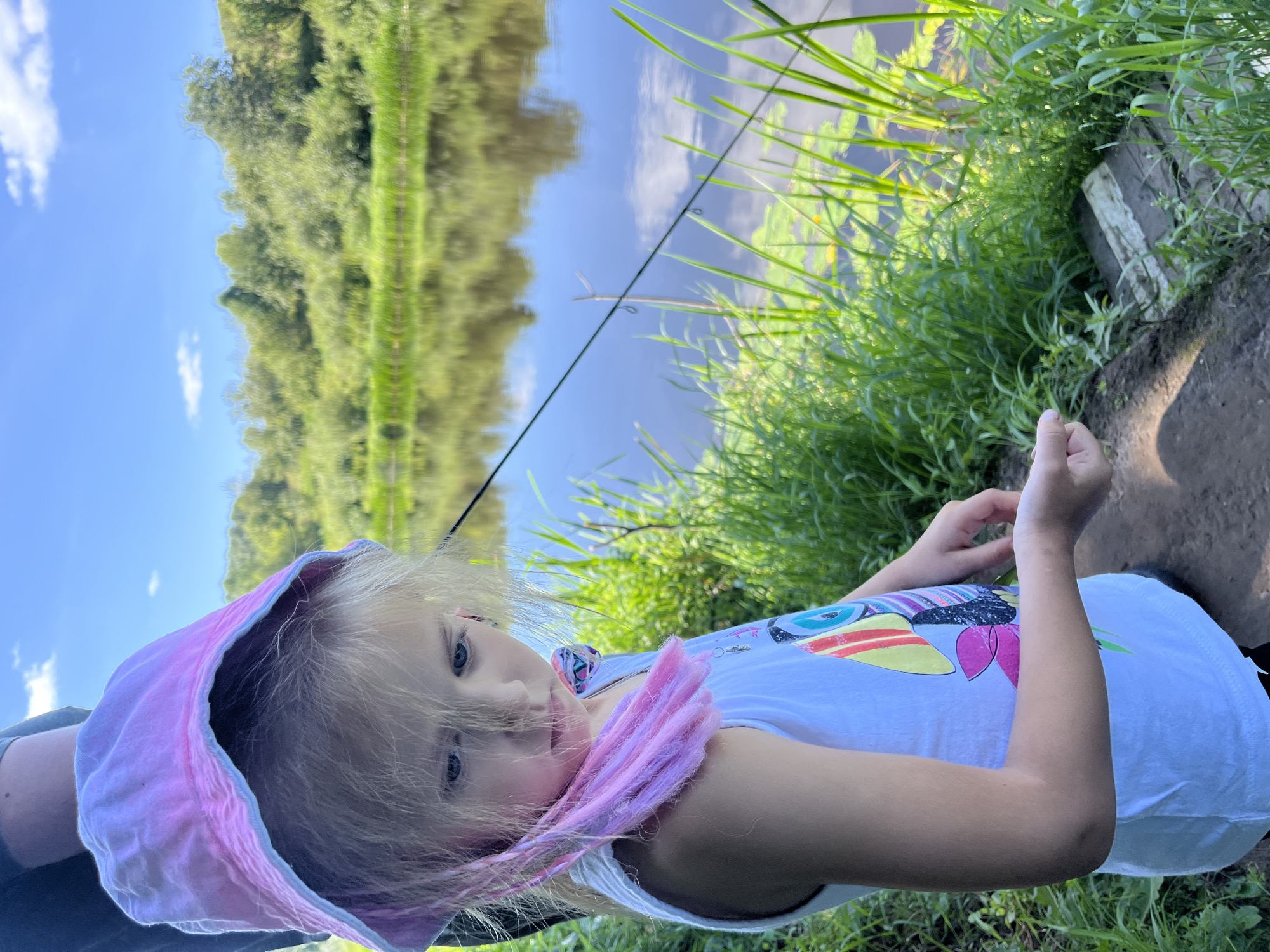 A young girl with blonde hair, wearing a pink sun hat and a white tank top with a colorful bird graphic, stands on a dirt path next to a river. Her gaze is directed downwards towards her left hand, which appears to be holding something small. The river reflects the blue sky and lush green trees and foliage on the opposite bank. In the foreground, tall green grass and lily pads are visible in the water. A fishing rod is also present, with its line extending across the water. The scene is set in a natural, outdoor environment, likely a rural area.