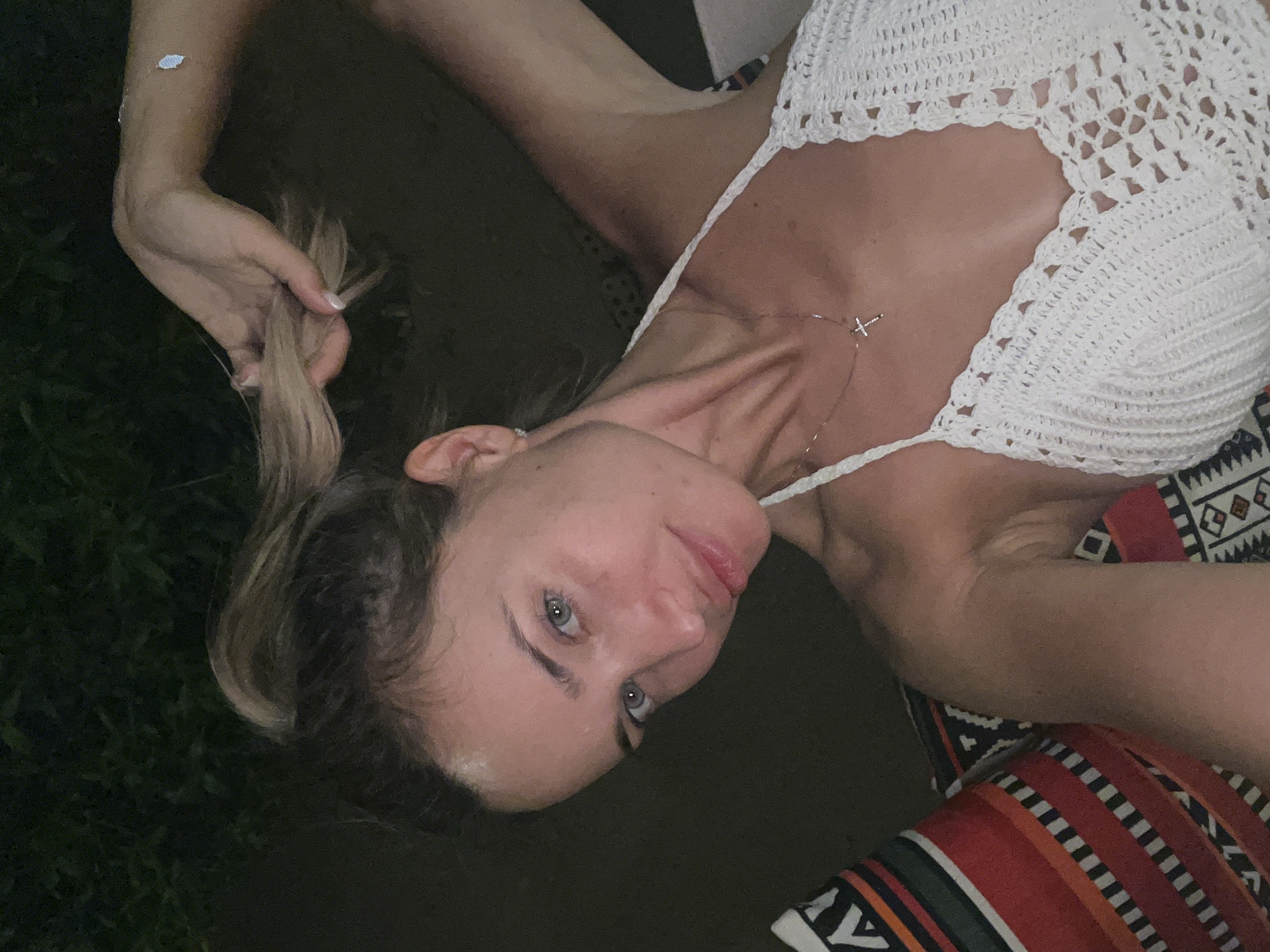 A woman with light skin, blue eyes, and blonde hair pulled into a ponytail is shown in a selfie taken at night. She is wearing a white crocheted top and a silver cross necklace. She is sitting on a patterned cushion. The background is dark, with greenery visible. This image was taken in Nuweiba, Egypt.