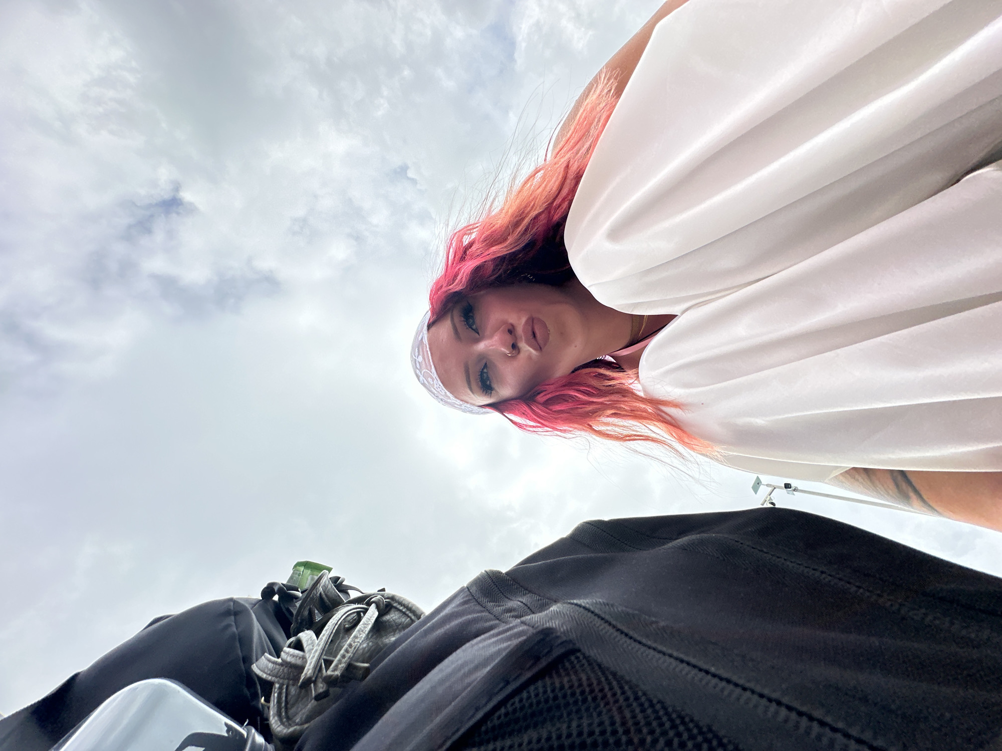 A low-angle shot shows a woman with pink hair, wearing a white top and headband, looking down at the camera. She has a nose piercing and light makeup. Behind her, a cloudy sky fills the background. In the foreground, a black bag or backpack is visible, along with some other items possibly strapped to it, including a water bottle. The location is unknown, but the prompt indicated the city of Kampung Toyapakeh, Indonesia. No specific actions or interactions are evident in the image.