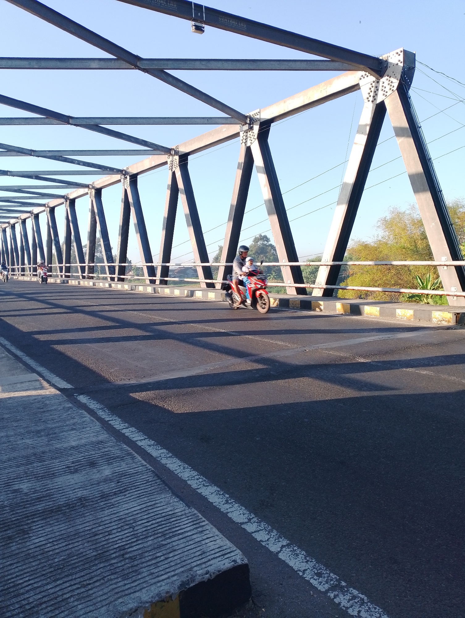 A wide-angle, eye-level outdoor shot captures a sunny day on a bridge in Mojo, Indonesia, with a focus on a motorcycle rider. The bridge's steel truss structure dominates the upper half of the image, casting stark geometric shadows across the asphalt road. The road surface itself appears worn, with visible white lane markings and a textured concrete section on the left.

In the mid-ground, a person wearing a helmet and a gray jacket rides a red motorcycle, with a passenger in the back. They are moving across the bridge, from left to right. Further in the distance, other figures on motorcycles are visible, suggesting traffic flow. The background reveals lush green foliage and trees under a clear blue sky. The lighting indicates a time of day when the sun is casting long shadows, likely late afternoon.