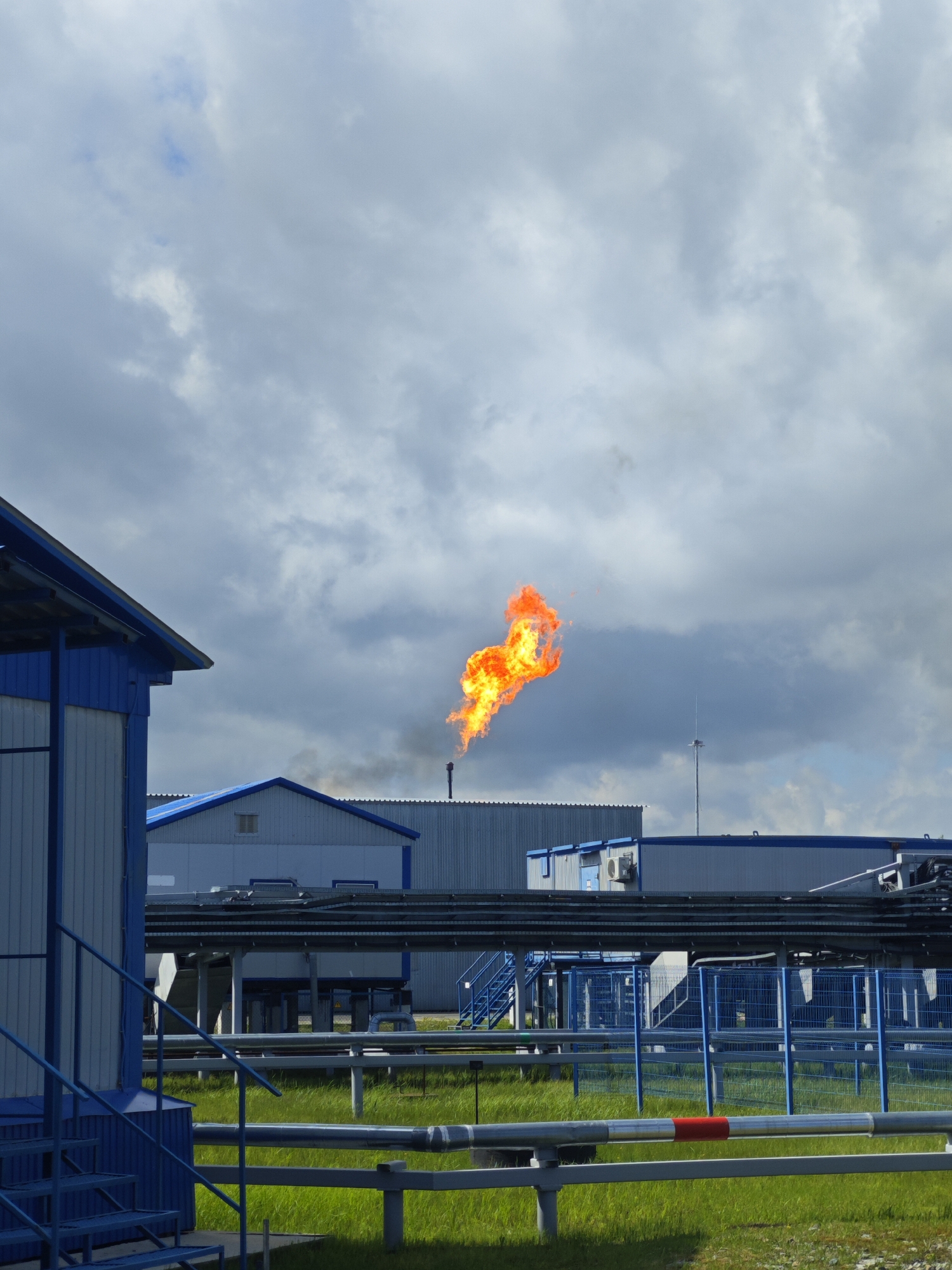 The image shows a gas flaring operation at an industrial site. A tall metal pipe is emitting a large flame of burning gas, creating a dark plume of smoke rising into the sky. The sky above is mostly cloudy with patches of blue. The industrial site contains several large buildings, painted in light blue and gray, with visible piping and infrastructure running between them. In the foreground, there's a smaller blue building with stairs. The ground is covered with green grass and metal fencing. The location appears to be an industrial area, possibly a gas processing or power generation facility.