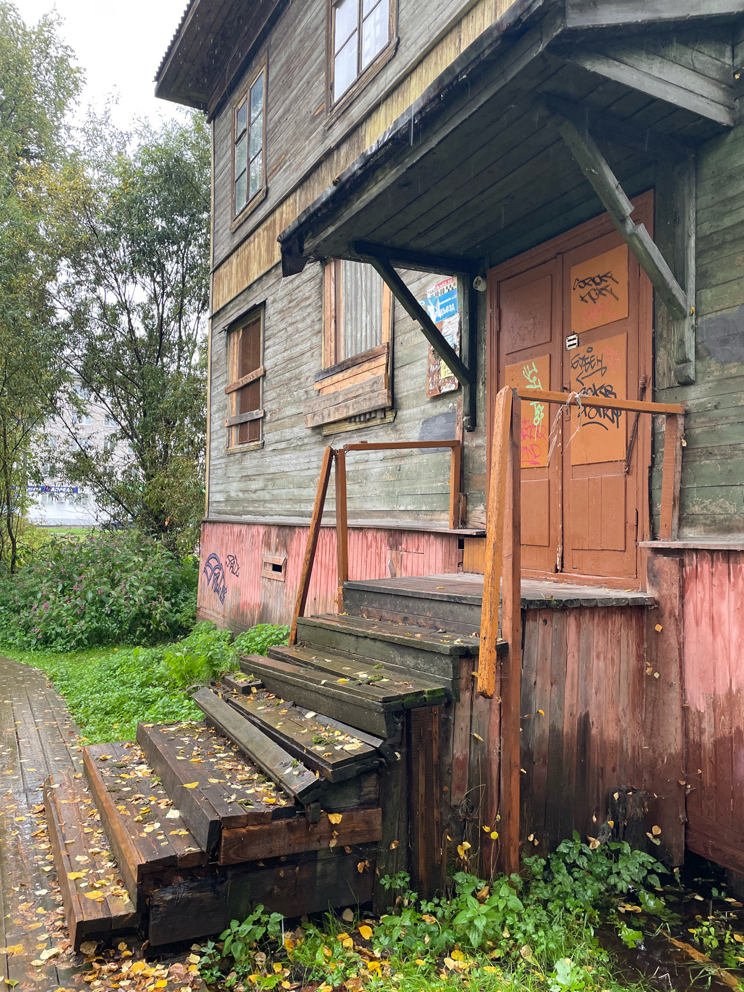 This image depicts an abandoned wooden apartment building, likely located in the city of Arkhangelsk, Russia. The building is weathered and shows signs of neglect, with peeling paint and graffiti on the doors and walls. The entrance features a dilapidated wooden staircase covered in fallen autumn leaves, with puddles of water at its base. The surrounding environment is overgrown with grass and bushes, indicating a lack of maintenance. The sky is overcast, suggesting a damp or rainy day. The overall impression is one of decay and abandonment.