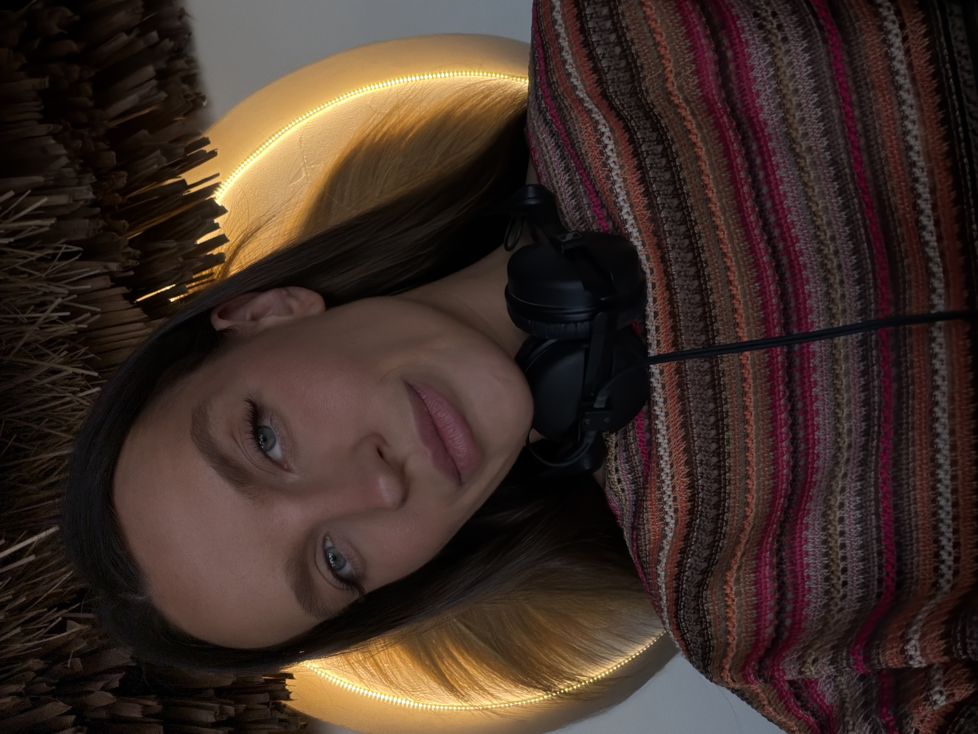 A young woman with dark hair and blue-green eyes is looking at the camera. She is wearing a colorful striped sweater and black headphones around her neck. The background is blurred, but it appears to be indoors with a circular light fixture and a thatched roof overhead. The setting is Drobnići, Montenegro.