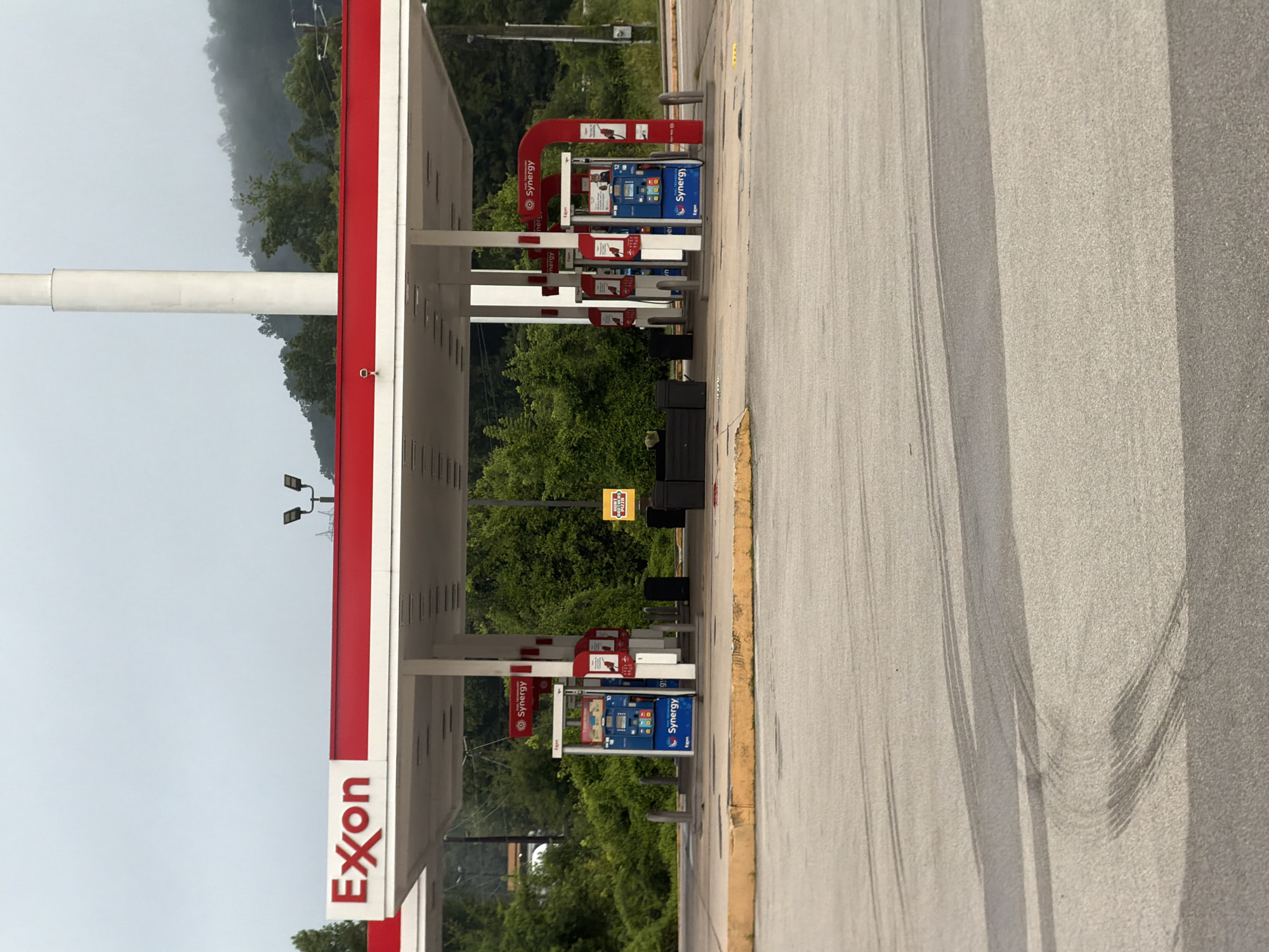 An exterior, medium shot captures an Exxon gas station located in Wildwood, United States. The scene is overcast with a foggy, mountainous backdrop. The gas station features a red and white canopy extending over gas pumps. Two blue and white Exxon Synergy gas pumps are visible on the left, while two red and white Synergy pumps are on the right. Trash cans are situated behind the pumps. A yellow sign with red lettering and the words "HONEST BROTHERS" is mounted on a pole to the right of the pumps. The asphalt in the foreground shows tire marks.