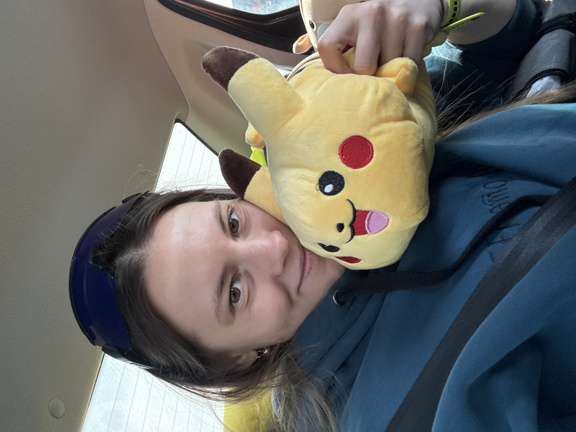 An image captures a young woman inside a vehicle, likely a car, holding two plush toys. She is positioned at an angle, looking directly at the viewer with a neutral to slightly smiling expression. Her dark brown hair frames her face, and she has brown eyes. A dark blue hoodie covers her, and a black safety belt diagonally crosses her chest. A dark blue headband or sunglasses are pushed up onto her head.
She cradles a yellow Pikachu plush toy against her left cheek, which features brown-tipped ears, red circular cheeks, black eyes, and a pink tongue peeking out. Behind Pikachu, a hand holds another, less visible plush toy, possibly a Fennekin, identifiable by its ear and fur color. A bright yellow or neon green wristband is visible on her right wrist.
The background shows the beige interior ceiling of the car and a window with vertical streaks, possibly indicating rain or condensation. Some unreadable white text is faintly visible on her hoodie. The scene suggests she is traveling, possibly as a passenger. No specific location cues beyond the car interior are discernible within the image itself for Mozhga, Russia. The lighting appears to be natural daytime light.