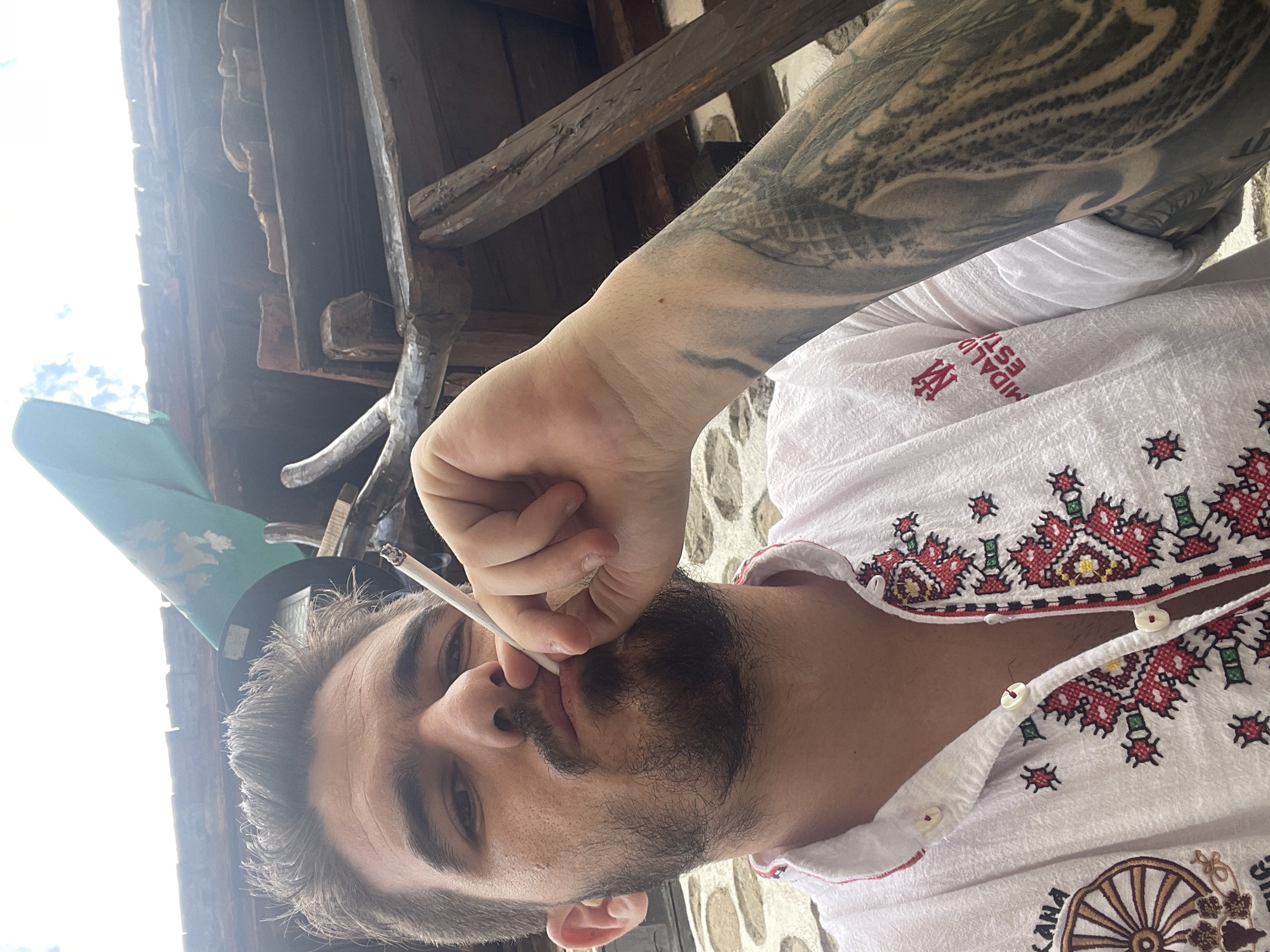 The image, taken from a low-angle perspective looking upwards, features a young man smoking a cigarette. He has a beard and mustache, with dark hair showing some grey strands. His gaze is directed slightly upwards and away from the camera as he holds a lit cigarette to his lips with his right hand.
He is wearing a white, short-sleeved shirt adorned with intricate red, green, and black traditional embroidery, concentrated around the collar and upper chest area. On the right chest of the shirt, the text "MIDALIDARE ESTATE" is visible next to a stylized logo resembling intertwined 'M' and 'E'. Further down the shirt, on the lower right, a circular, wheel-like design is embroidered alongside the text "KANA". The man's right arm is heavily tattooed with dark, full-sleeve patterns that appear abstract or tribal.
The setting suggests an outdoor or semi-outdoor location during bright daytime, with a clear sky partially visible through a rustic overhead wooden beam structure. This wooden framework, possibly part of a veranda or an old building's ceiling, is prominent in the upper background. Behind the man's right arm and shoulder, a textured surface resembling a stone wall or paving is visible. A corner of a green, conical object, possibly a party hat or a flag, is seen behind his head on the left side of the frame. The man's expression is neutral and calm.
