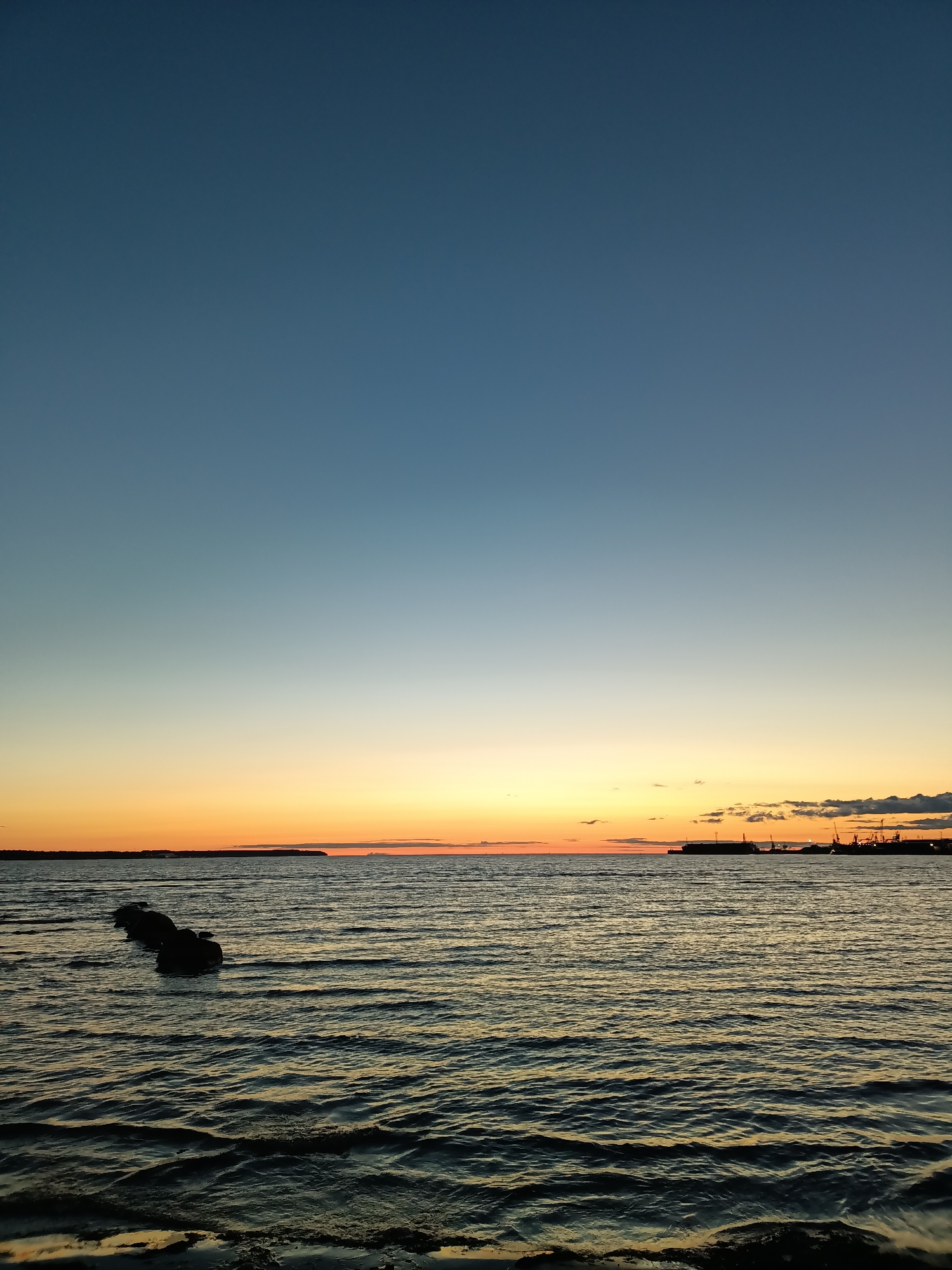 The image captures a serene sunset over the Baltic Sea, as seen from the coast of Tallinn, Estonia. The sky transitions from a deep blue at the top to hues of orange and yellow near the horizon, indicating the sun has recently set or is about to rise. The sea is calm, with gentle waves rippling across its surface, reflecting the colors of the sky. In the foreground, a line of dark, submerged objects, possibly remnants of an old pier or breakwater, breaks the water's surface. In the distance, the silhouette of the Tallinn port is visible, with cranes and ships creating a stark contrast against the colorful sky. The overall scene evokes a sense of tranquility and natural beauty, characteristic of coastal sunsets.
