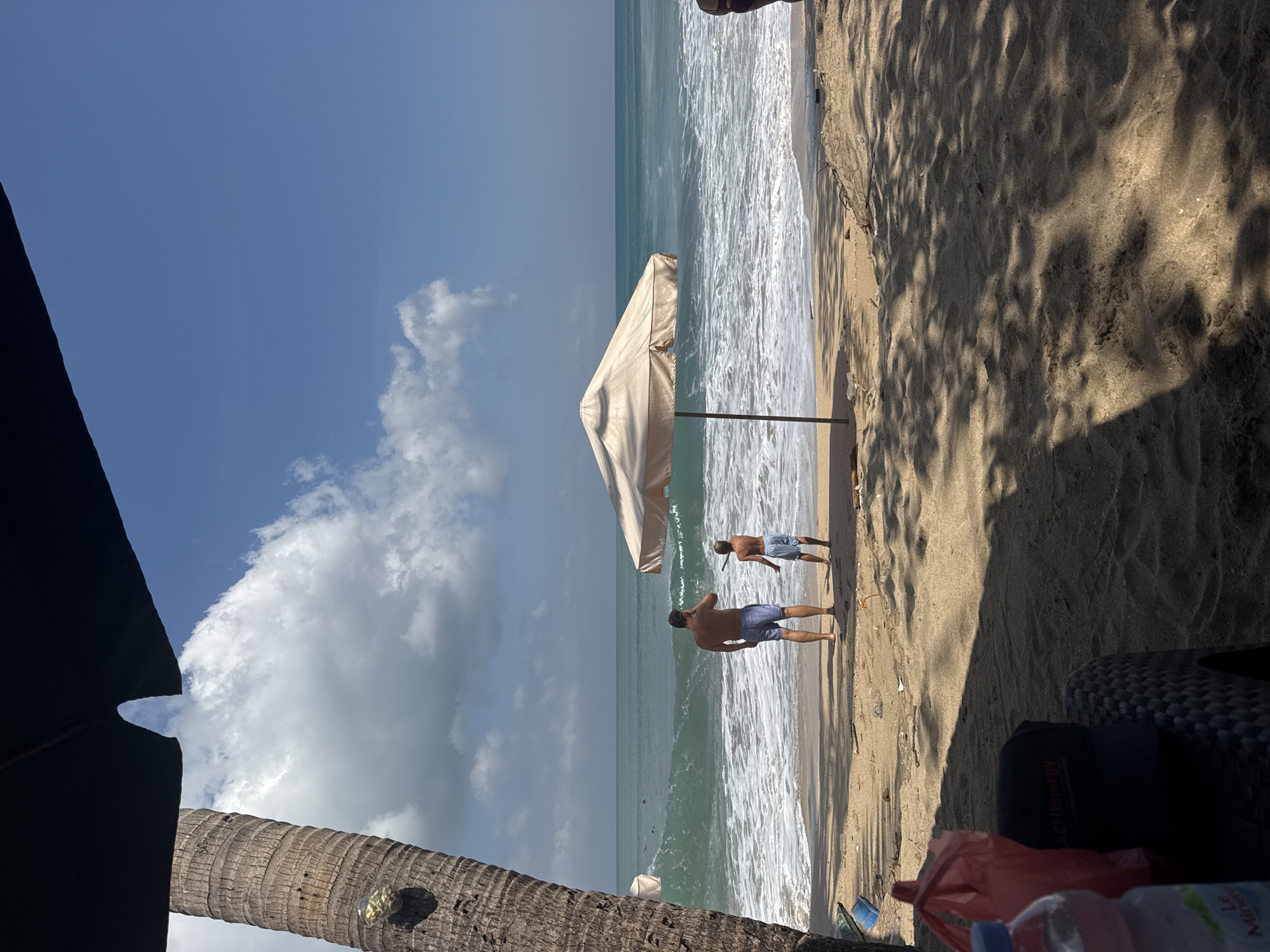 In Kuta, Indonesia, two individuals stand on a sandy beach facing the ocean. The person on the left, a man, is wearing blue patterned shorts. To his right, a boy, also in blue shorts, holds a stick. Both are looking towards the water where waves are breaking. A light-colored beach umbrella stands between them and the ocean. In the foreground, a large palm tree trunk is visible on the left, and shadows from foliage are cast across the sand. A plastic bottle and red plastic bags are on a black woven surface in the lower left corner. The sky is blue with scattered white clouds. The ocean water is a turquoise green.