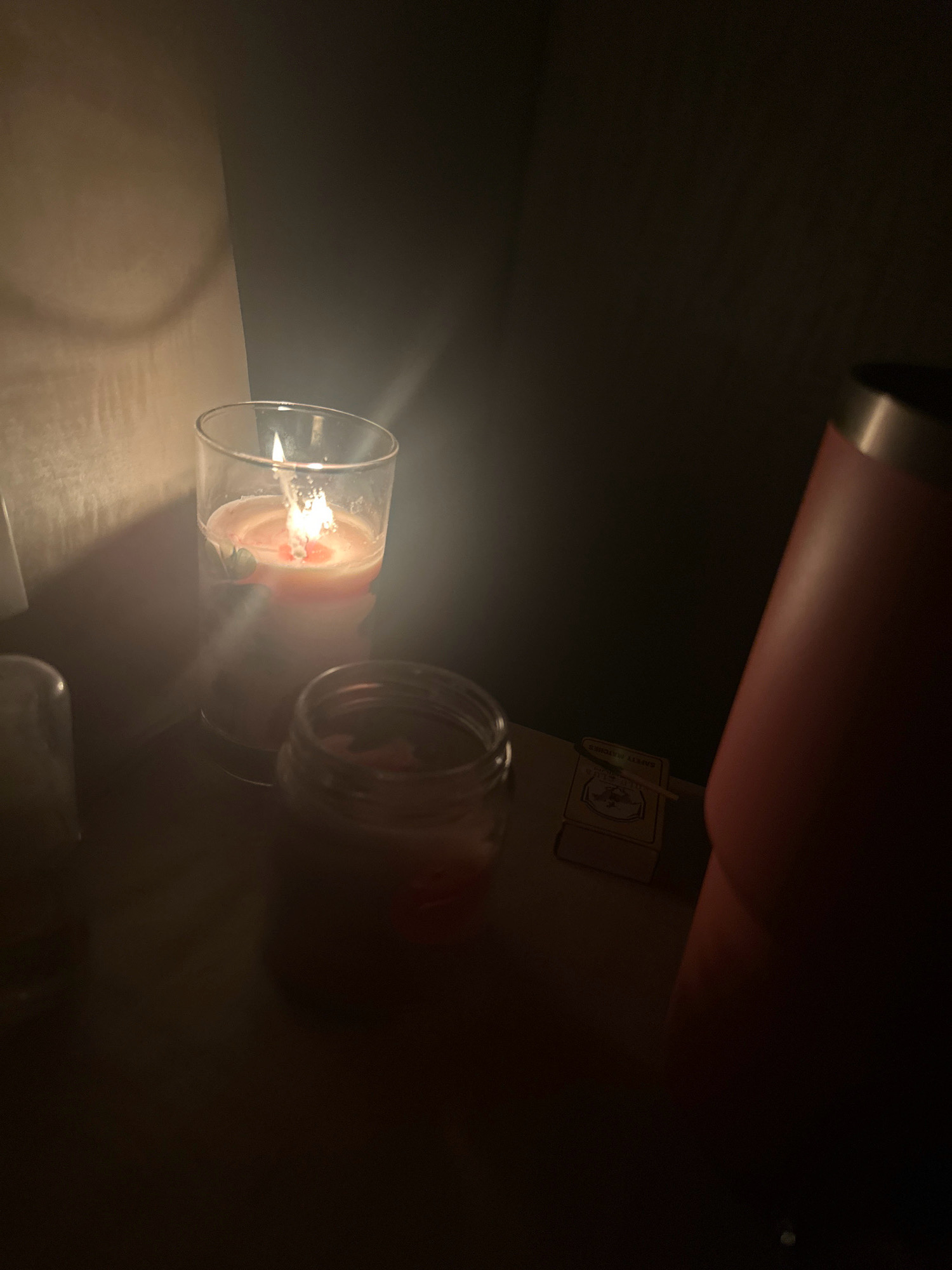 An indoor scene in Lalitpur, Nepal, features a lit candle in a glass jar, casting a warm glow on the surrounding objects. The candle is positioned on a flat surface, likely a table or shelf, and appears to be made of wax with a visible wick. Next to the candle, a small box of matches is visible, suggesting the candle was recently lit. The background is dimly lit, with shadows creating an atmospheric effect. The overall impression is one of a peaceful and cozy setting, possibly a home or a small establishment in the city.