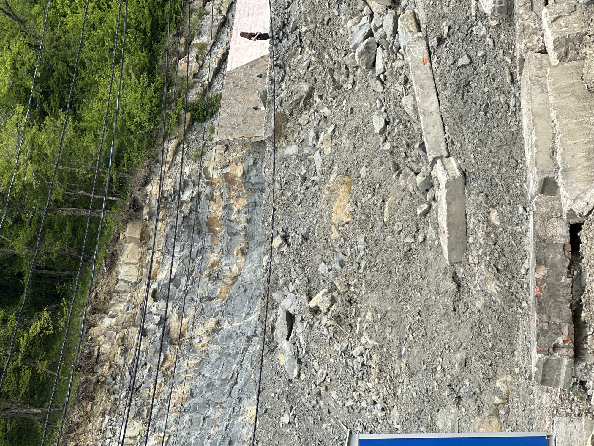 The image shows a steep, rocky hillside next to a road or path in Novomikhaylovsky, Russia. The hillside consists of exposed layers of gray and tan rock. Power lines run along the edge of the hillside, near the trees. Debris, rocks, and what appears to be broken concrete are scattered on the ground. A woman is visible standing near the top edge of the rocky area. The image is taken from an elevated perspective, looking down onto the hillside. The sky is not visible.