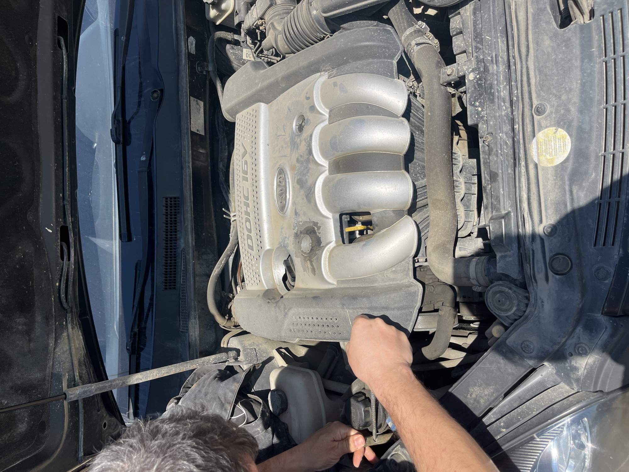 The image depicts a person working on the engine of a car. The engine bay is visible, with various engine components such as the air intake manifold and coolant reservoir in view. The person is likely performing maintenance or repairs on the vehicle. The make and model of the car is Kia. The location is not identifiable from the image.