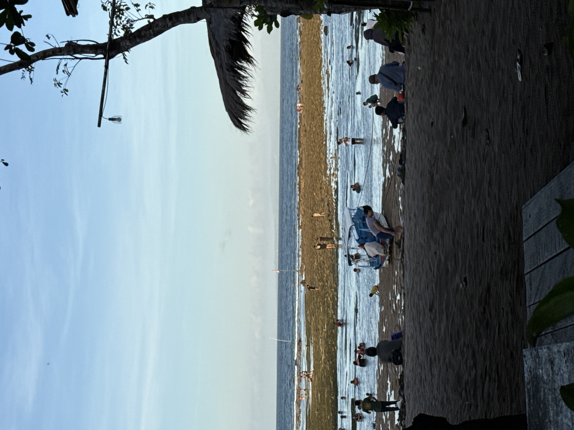 The image depicts a beach scene, possibly in the city of Denpasar, Indonesia. A sandy beach transitions into a shallow, light-colored body of water with people wading and interacting. The sky is light blue with a few clouds. A tree with a light fixture hangs over the scene on the left side of the image. The photo has been rotated 90 degrees clockwise.