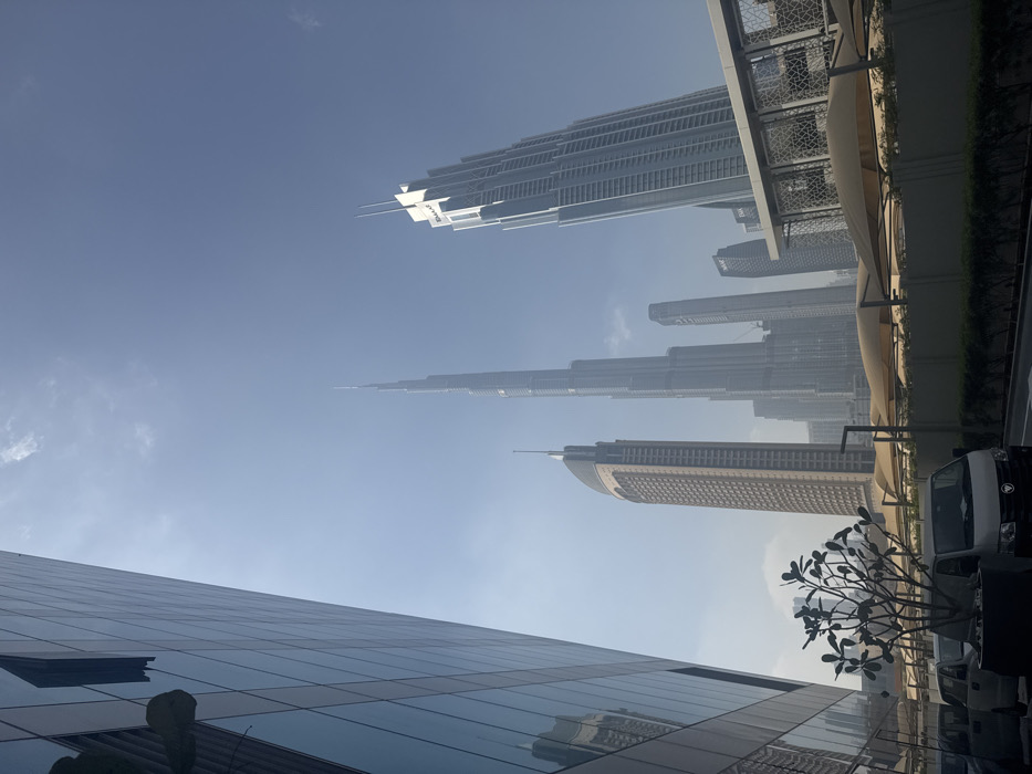 Dubai's skyline is depicted from a low angle, emphasizing its towering skyscrapers against a hazy blue sky. The Burj Khalifa dominates the view, flanked by other high-rise buildings. Reflections of structures are visible in the glass facade of a building in the foreground. Part of a parking area is shown with cars and a small tree adding a sense of scale to the image. The image is rotated to the side.