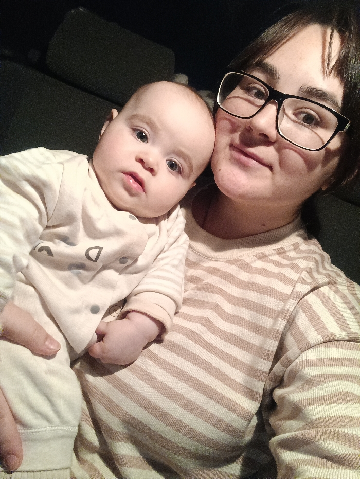 A close-up, warm-toned selfie shows a woman holding a baby in what appears to be an indoor or dimly lit setting, possibly a car interior given the dark, textured background resembling upholstery.
On the right side of the frame, a woman with dark, short hair and black-framed glasses (with a white inner edge) looks directly at the camera with a gentle, soft smile. She is wearing a long-sleeved shirt with horizontal stripes in alternating shades of beige/light brown and off-white.
Cradled in the woman's arms, on the left side of the frame, is a baby looking intently at the camera with wide, curious dark eyes and a neutral expression. The baby has light, fine hair and chubby cheeks. The baby is dressed in a light-colored, possibly off-white or very pale beige, full-body outfit or sleepsuit adorned with faint grey, abstract shapes or patterns (such as a 'D' shape and circles). The baby's tiny hand is visible near the woman's chest.
The lighting is soft and somewhat muted, casting gentle shadows and highlighting the faces of the woman and baby. The overall impression is one of affection and connection between the two individuals.