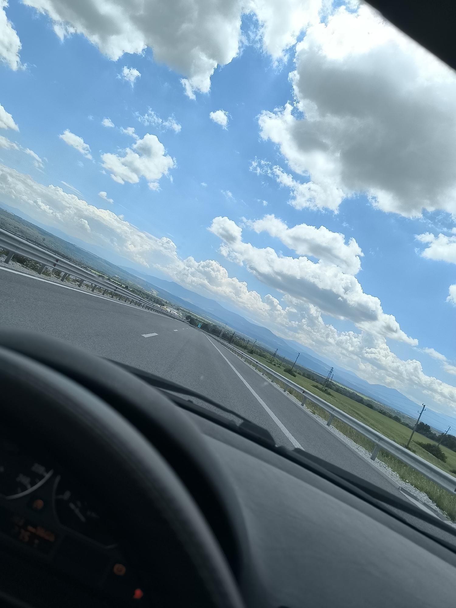 The image depicts a highway scene near Valchek, Bulgaria, captured from the interior of a vehicle. The upper portion of the frame features a bright blue sky filled with numerous white cumulus clouds. The lower foreground presents the upper section of a dark-colored steering wheel and a segment of the vehicle's dashboard. Visible within the instrument cluster are an illuminated red warning light, an amber warning indicator, and a digital readout displaying '75' alongside other alphanumeric characters. The multi-lane asphalt highway, marked with white lane lines, extends into the distant horizon. Metal guardrails line both sides of the roadway. Beyond the guardrails, green fields transition into a landscape of distant mountains under the cloudy sky. Utility poles with power lines are discernible along the right side of the highway. No individuals are visible within the scene.