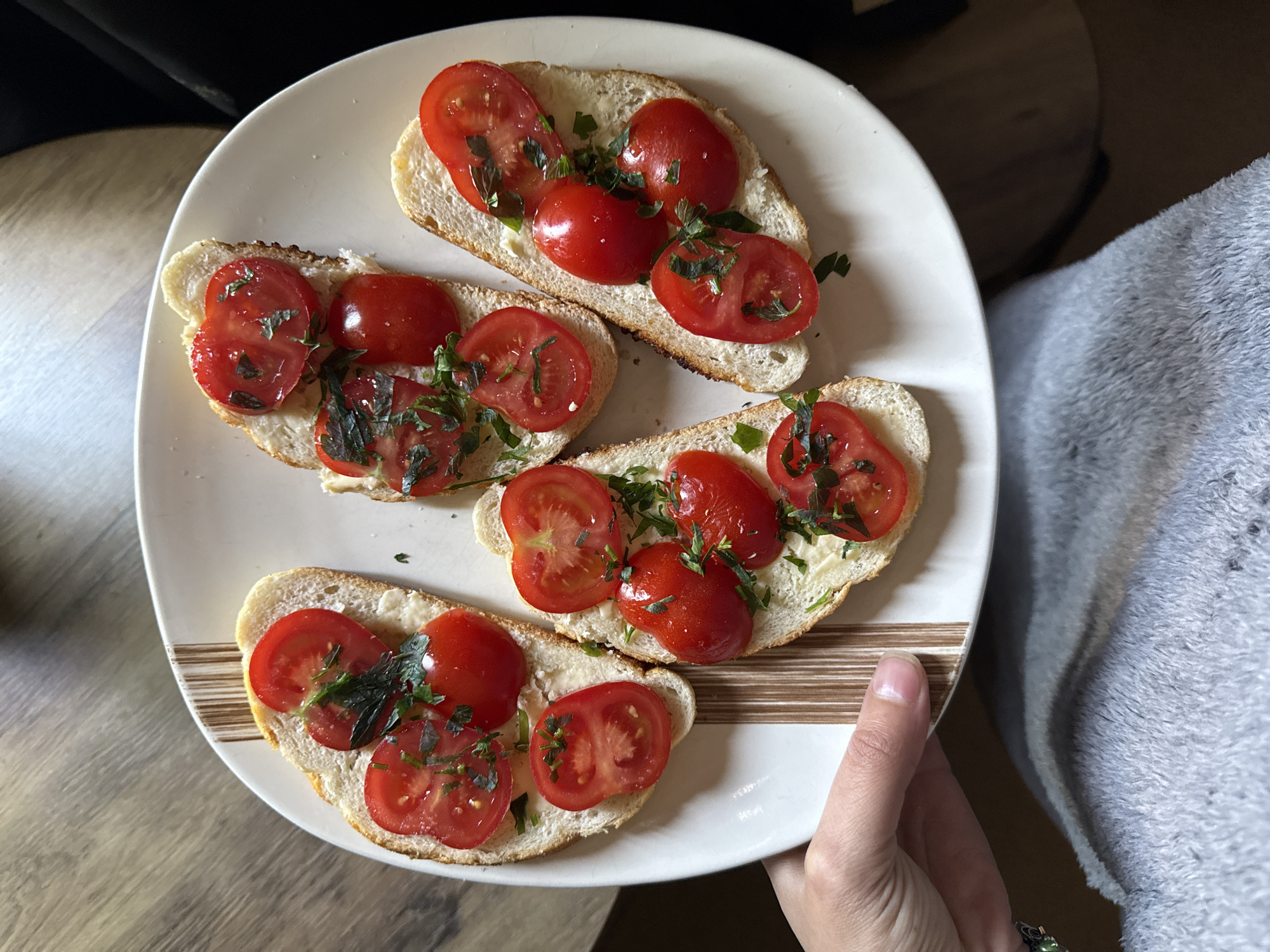 A close-up, high-angle shot features a person's hand holding a white ceramic plate laden with four individual slices of bread topped with tomatoes and herbs. The bread appears to be a type of baguette or similar crusty loaf, lightly toasted or fresh, with a visible white spread underneath the toppings, possibly butter or cream cheese. Each slice is garnished with bright red tomatoes, including some whole cherry tomatoes and others neatly halved, providing a vibrant contrast against the white bread. A generous sprinkle of chopped fresh green herbs, likely basil or parsley, adds a finishing touch and color.
The human hand, positioned on the bottom right of the frame, has fair skin and short, natural fingernails. A silver ring adorned with a distinct green gemstone, possibly emerald or jade, is visible on the ring finger, adding a touch of personal detail.
The plate itself has a decorative brown striped pattern along its rim. The background on the left reveals a light-colored wooden surface, possibly a table, exhibiting a subtle wood grain. To the right of the hand, a soft, grey, textured fabric, resembling a blanket or warm clothing, is visible. The scene is set indoors, likely in a domestic environment such as a kitchen or dining area, indicated by the home-style food presentation and background elements. The lighting suggests it is daytime. The food, resembling bruschetta, appears ready to be served or eaten. No text is visible within the image.