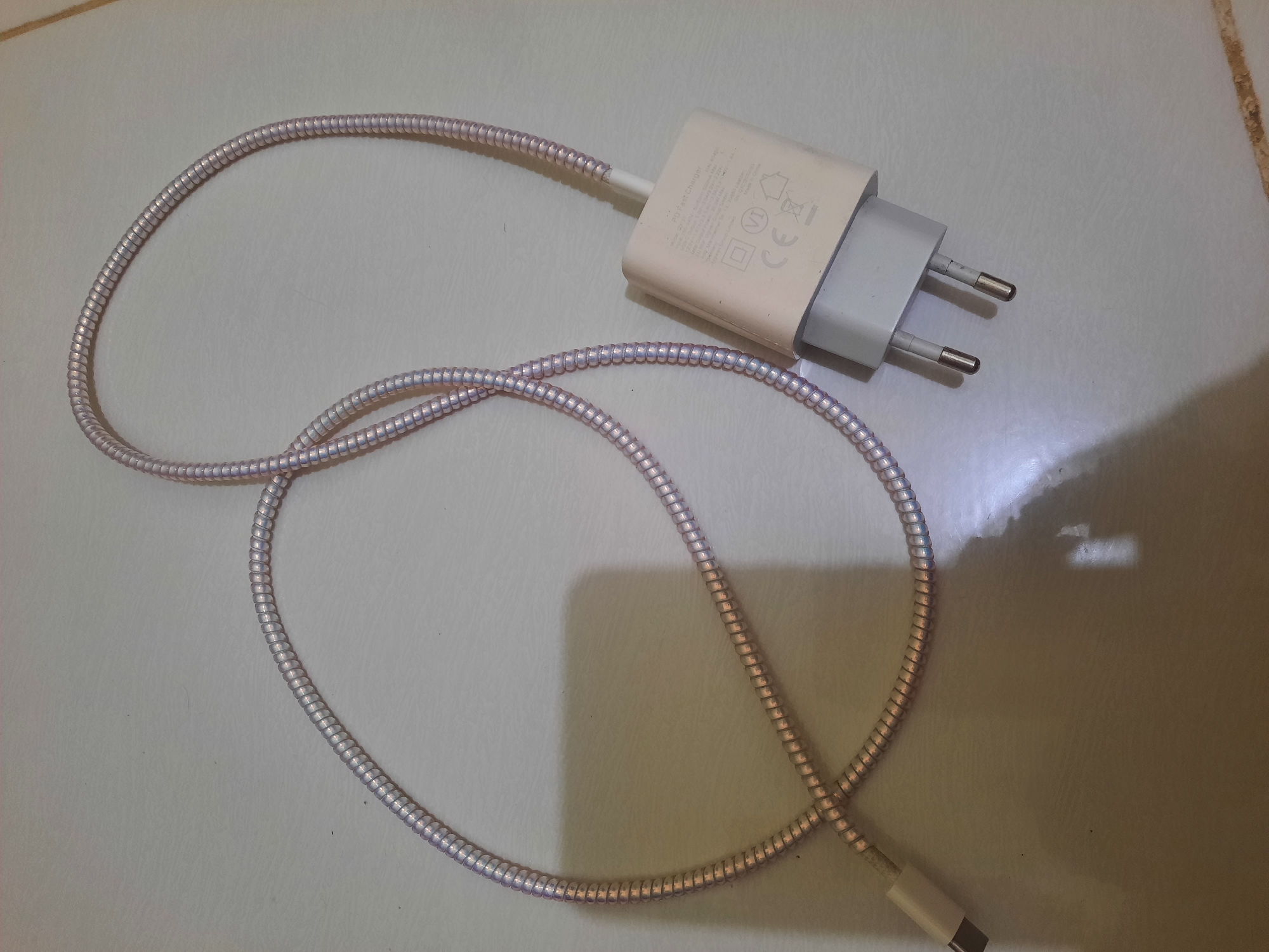 This is a top-down, medium shot of a white and grey USB-C charger with a braided silver cable. The charger has two prongs for a European-style outlet. The cable is coiled in a loop and extends to the left, where a USB-C connector is visible. The charger has text printed on it, but it is too small to read clearly in this image. The charger and cable are lying on a light-colored, tiled surface that appears to be a floor. There is a shadow cast on the surface to the left of the charger, suggesting overhead lighting. No people are visible in the image. The scene appears to be indoors. The presence of European-style electrical plugs suggests the charger might be used in regions with that standard. No specific details about Lhokseumawe, Indonesia are discernible from the image itself.