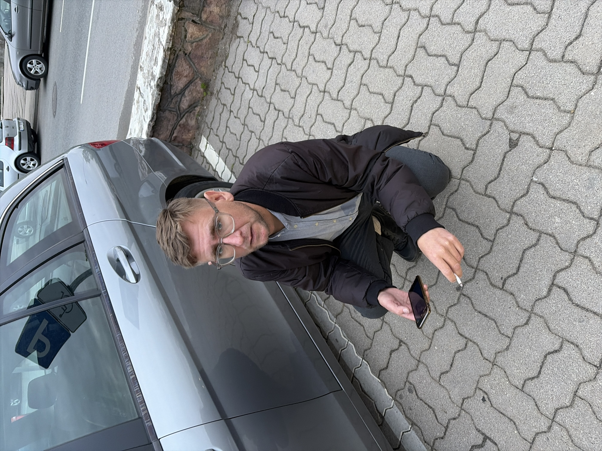 A man with glasses and light brown hair is kneeling on a paved area next to a grey car. He is wearing a dark jacket over a light blue collared shirt and dark pants. The man is holding a lit cigarette in his right hand and a smartphone in his left hand, looking towards the camera. The scene appears to be an outdoor parking lot, with other cars and a stone wall visible in the background. The paving stones are interlocking and grey. A sign with a wheelchair symbol is reflected in the car window. The image is taken from a high angle, looking down at the man. The lighting suggests it is daytime. There is no visible text other than the reflection on the car window.