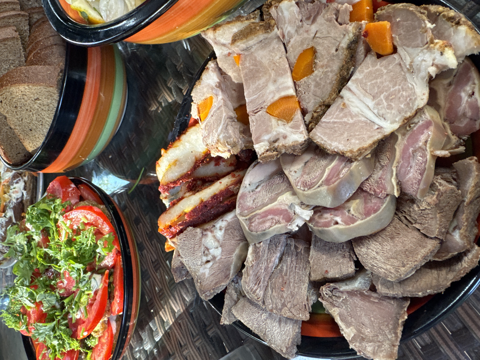 This is a close-up, overhead shot of a spread of food on a dark, possibly glass or reflective, table, set outdoors in what appears to be Sary-Oy, Kyrgyzstan, given the context. In the foreground, a black platter is filled with various sliced meats. Some pieces are dark brown and appear to be cooked muscle meat, while others are lighter pink and white, possibly including fat or different types of cuts like tongue or pork belly. There are also some pieces that have a reddish spice coating. Scattered amongst the meat are bright orange pieces, likely carrots.
Behind the platter of meat, to the left, is a colorful, striped ceramic bowl filled with a tomato and herb salad. The tomatoes are sliced and mixed with what looks like fresh green herbs, possibly parsley or dill, and a white dressing. Further back, to the right, is another colorful, striped ceramic bowl filled with slices of dark bread. Beyond these, other dishes are partially visible, suggesting a larger meal. The lighting indicates it is daytime, with sunlight casting shadows on the table. There are no people visible in the image.