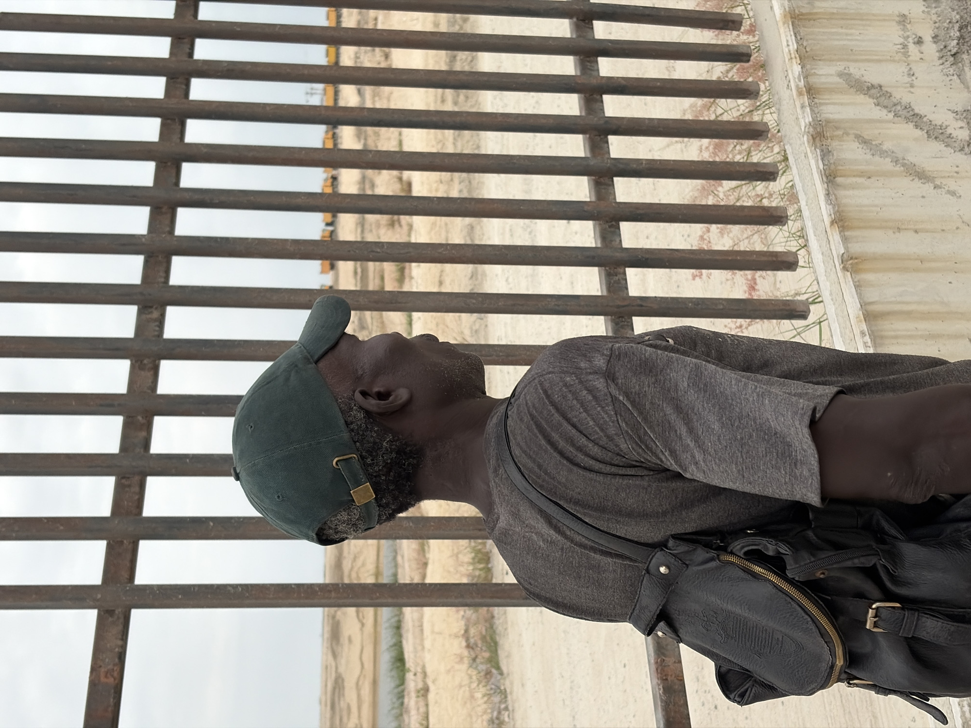 A medium shot captures a Black man wearing a dark green baseball cap and a dark gray t-shirt, with a black backpack slung over his left shoulder. He is positioned facing to the right, his gaze directed outwards, obscuring his eyes from view. The man's skin is dark, and his hair is short and curly. His expression is neutral.
The man stands in front of a tall, dark metal fence. The fence is made of vertical bars, spaced relatively close together. Beyond the fence, the landscape appears arid, with patches of dry grass and sand. In the distance, a train with multiple yellow cars is visible on a track. The sky is overcast and light gray.
The foreground shows the top of a concrete wall, which has a rough texture and is stained with brown streaks. The overall impression is that of an outdoor setting, possibly on the outskirts of a populated area, given the presence of the train. The specific location in the city of Itirin, Nigeria is not explicitly indicated by any visual cues in the image.