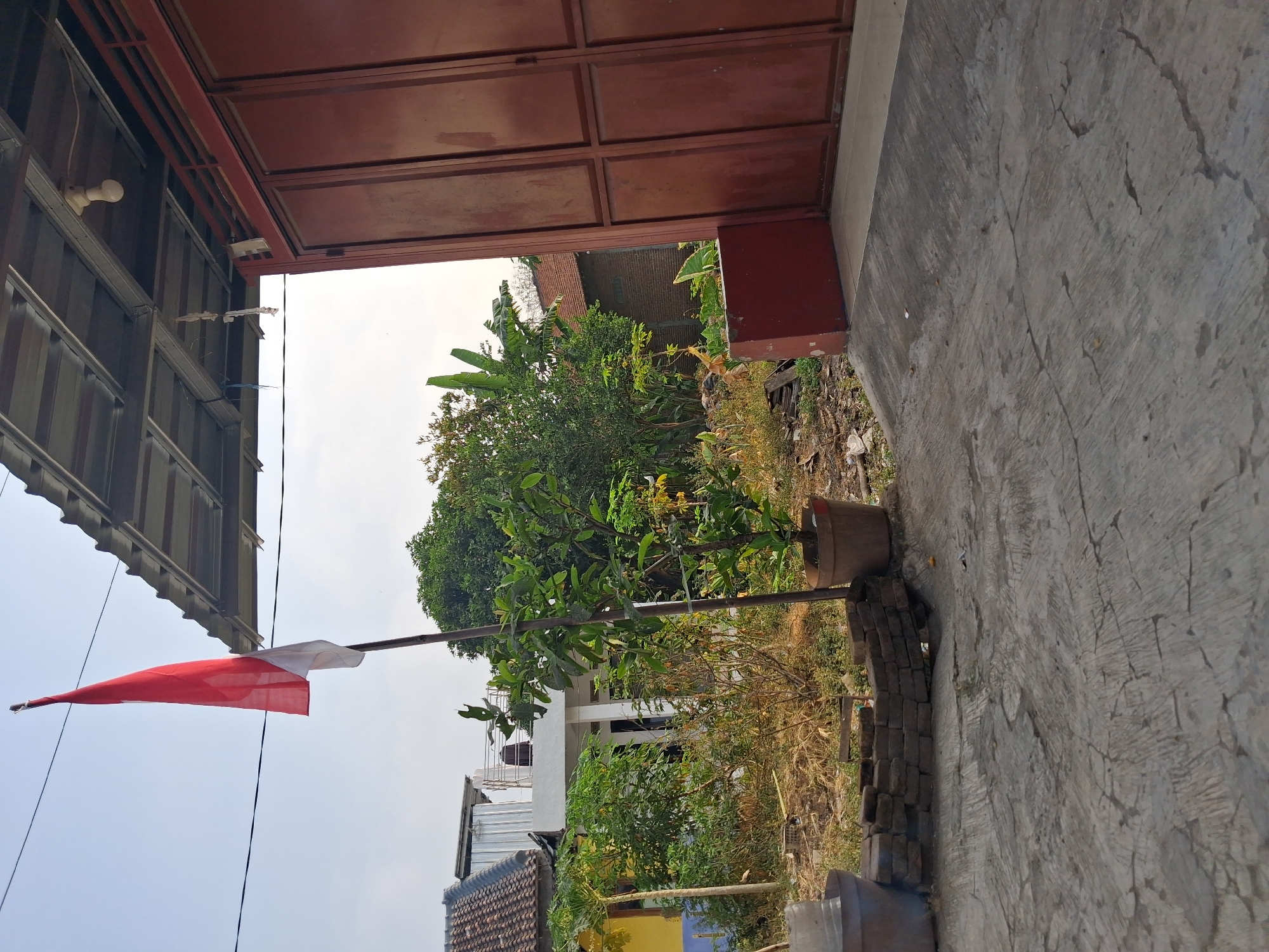 The image captures an outdoor scene in Balapulang Wetan, Indonesia, featuring a red and white Indonesian flag on a flagpole. The flag is positioned in front of a residential area, with houses and trees visible in the background. To the right, a large, reddish-brown metal gate and a section of a building's roof with visible metal framing and a light bulb are present. In the foreground, a cracked concrete surface leads towards the gate. There are various plants and some debris scattered in the mid-ground, including bricks and terracotta pots. The sky is clear and bright, suggesting daylight. No people are visible in the image.