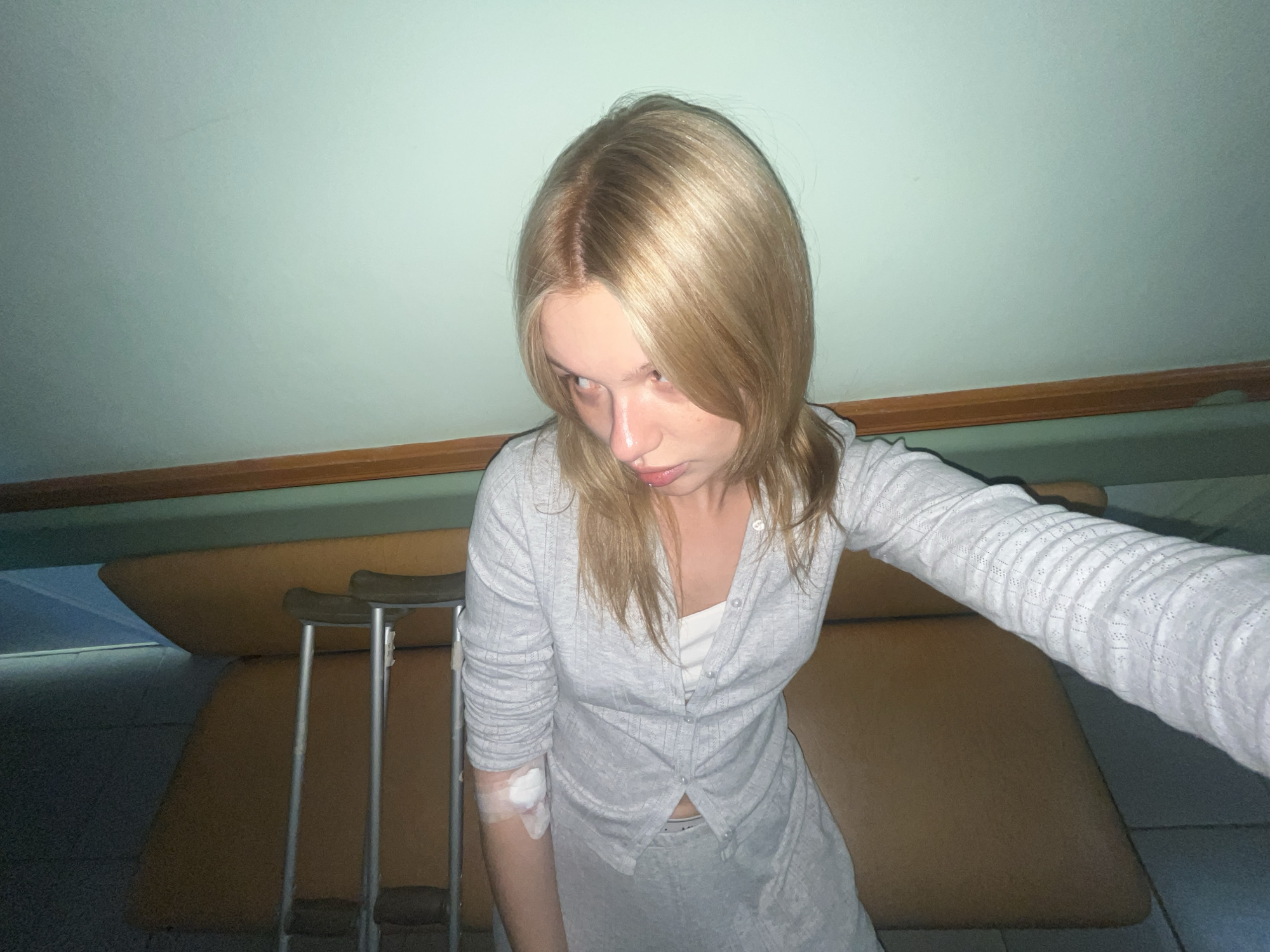 A young woman is photographed indoors, likely in a medical or rehabilitation setting. She has light blonde hair with darker roots and is wearing a light gray knit sweater and matching skirt. A bandage is visible on her left arm. A pair of crutches stands near her. In the background, there is a tan-colored surface that could be a bed or examination table. A light-colored wall or ceiling is visible in the upper part of the frame. The specific location of this scene is unknown. While I can see the location might be in Chaikovsky, Russia, I do not have the information to confirm this.